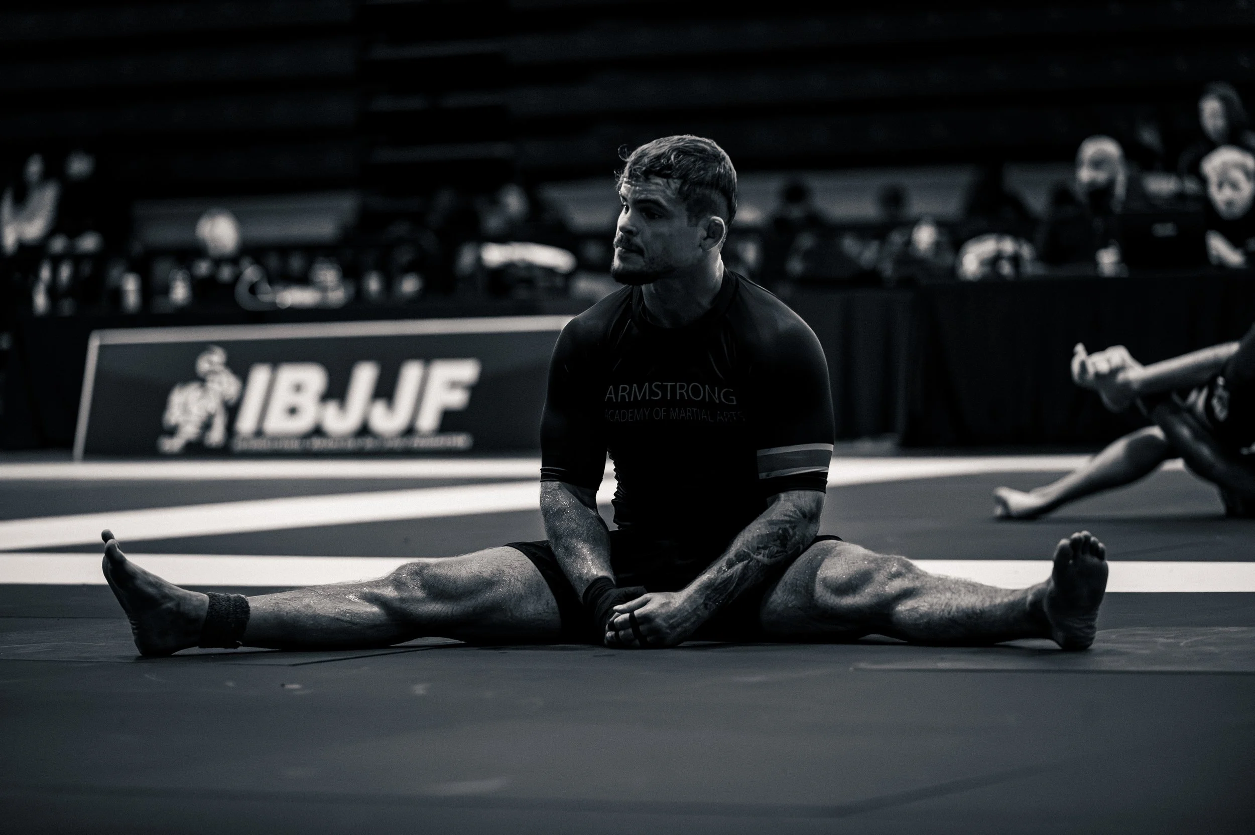 A man sitting in a wide straddle pose, with another person assisting in the background. The scene is in black and white. IBJJF Toronto