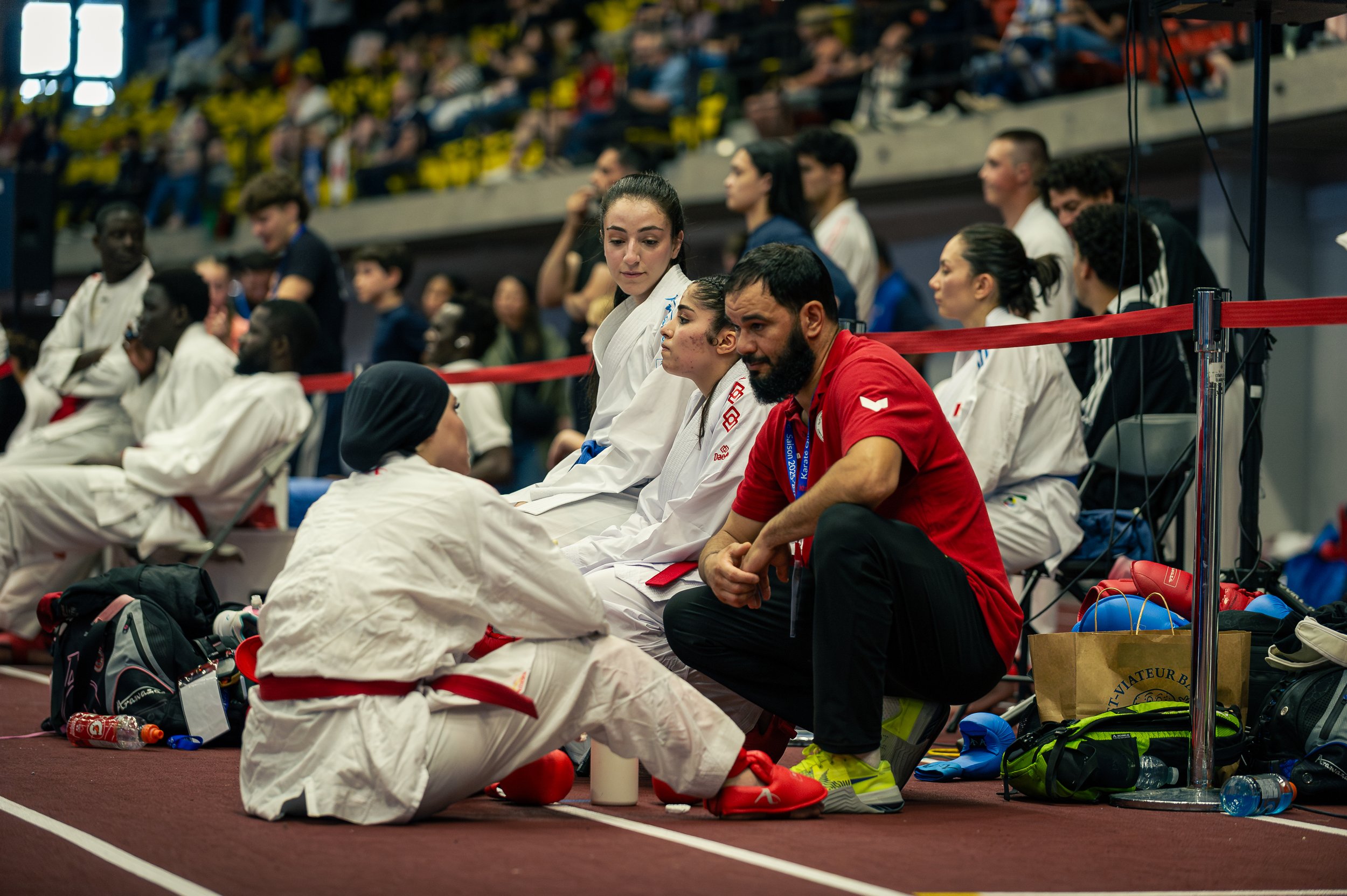 Athletes and coaches sitting on the sideline of an indoor sports arena during a martial arts event, with competitors in gi uniforms and various sports bags and equipment around them.
