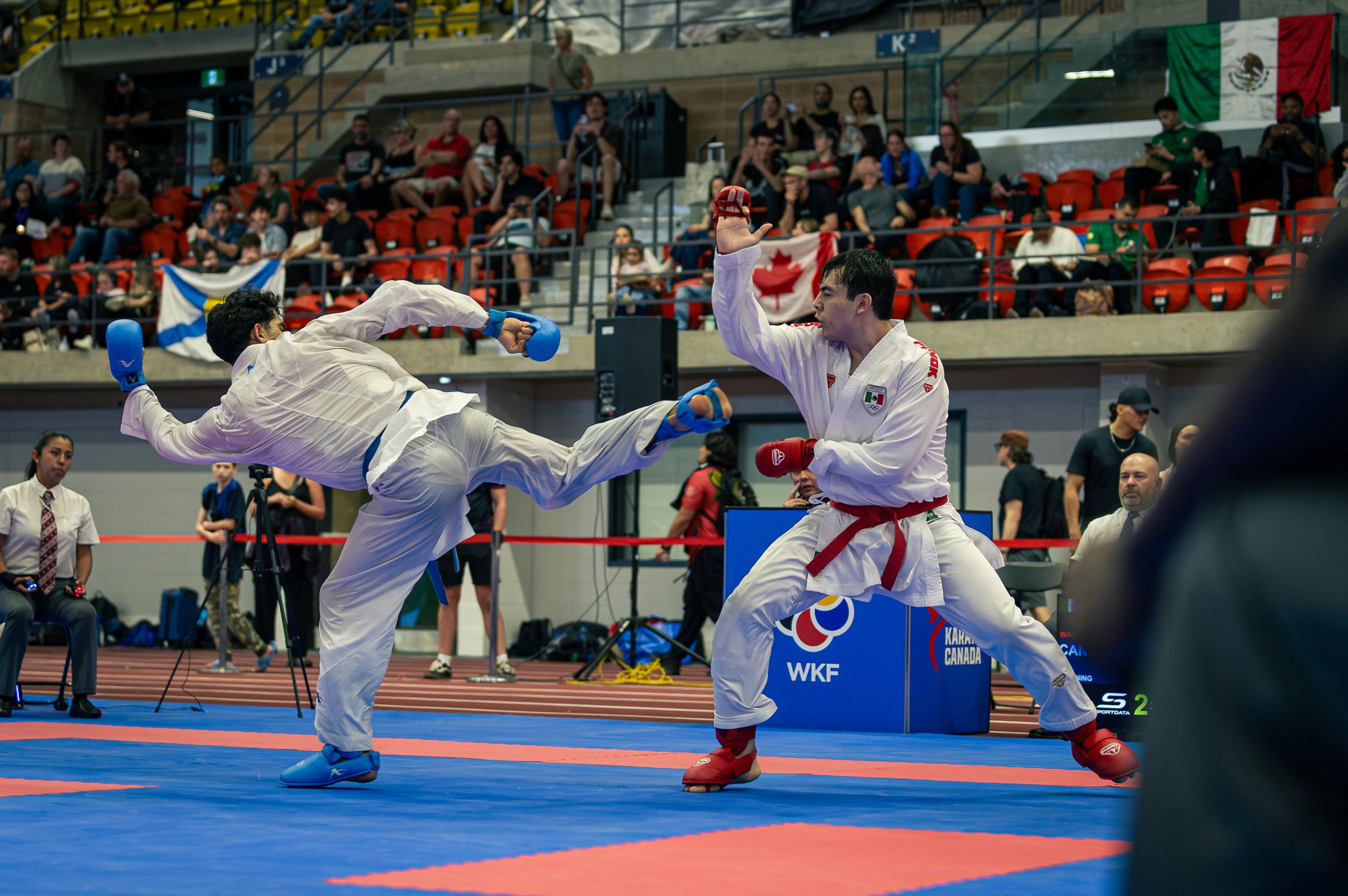 Two martial artists competing in a karate match on a blue and red mat at an indoor arena with spectators and flags, including the Mexican flag, in the background.