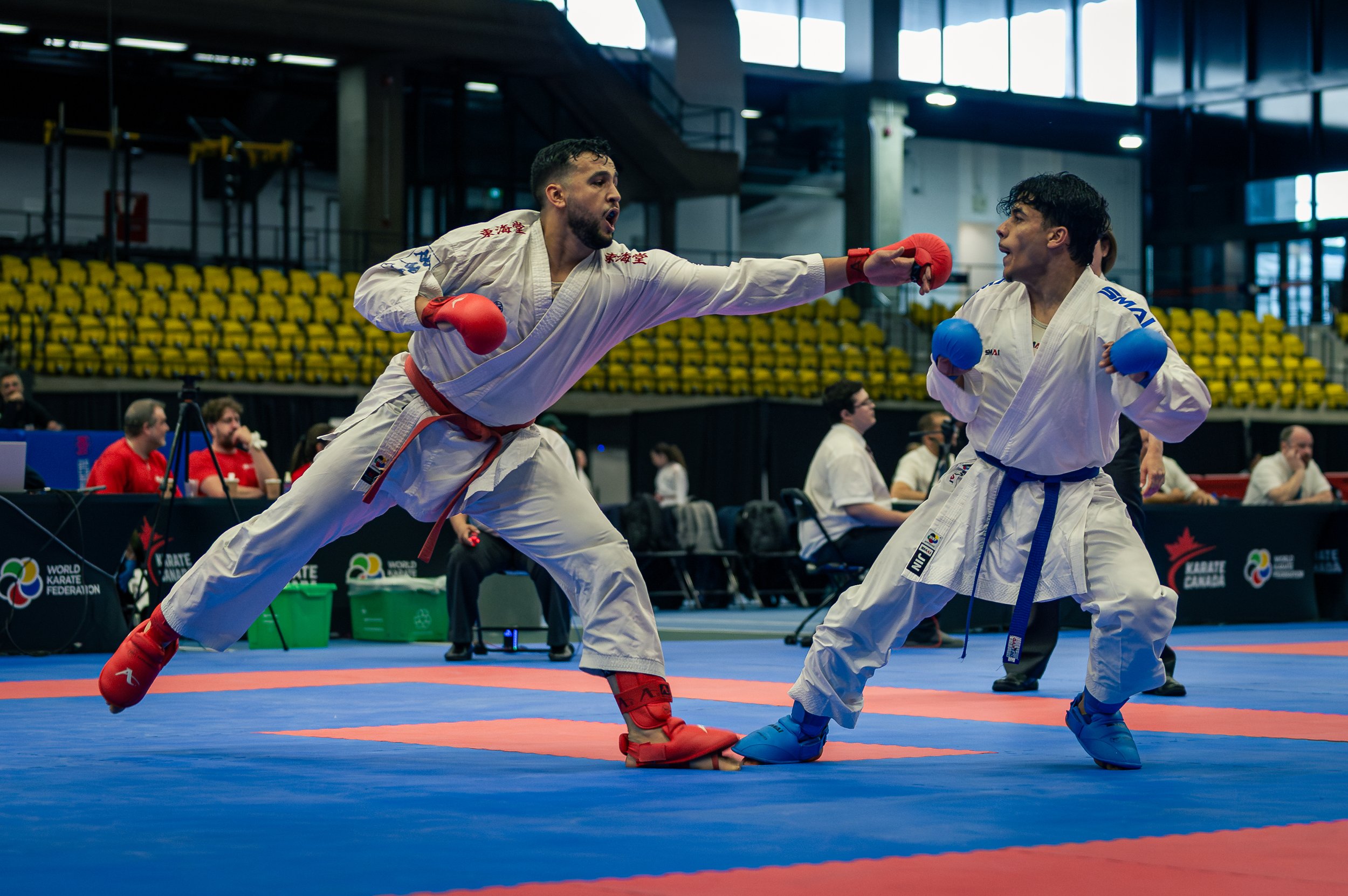 Two martial artists competing in a karate open international, one delivering a punch while the other blocks, in a centre claude robillard with yellow seats and onlookers in the background.