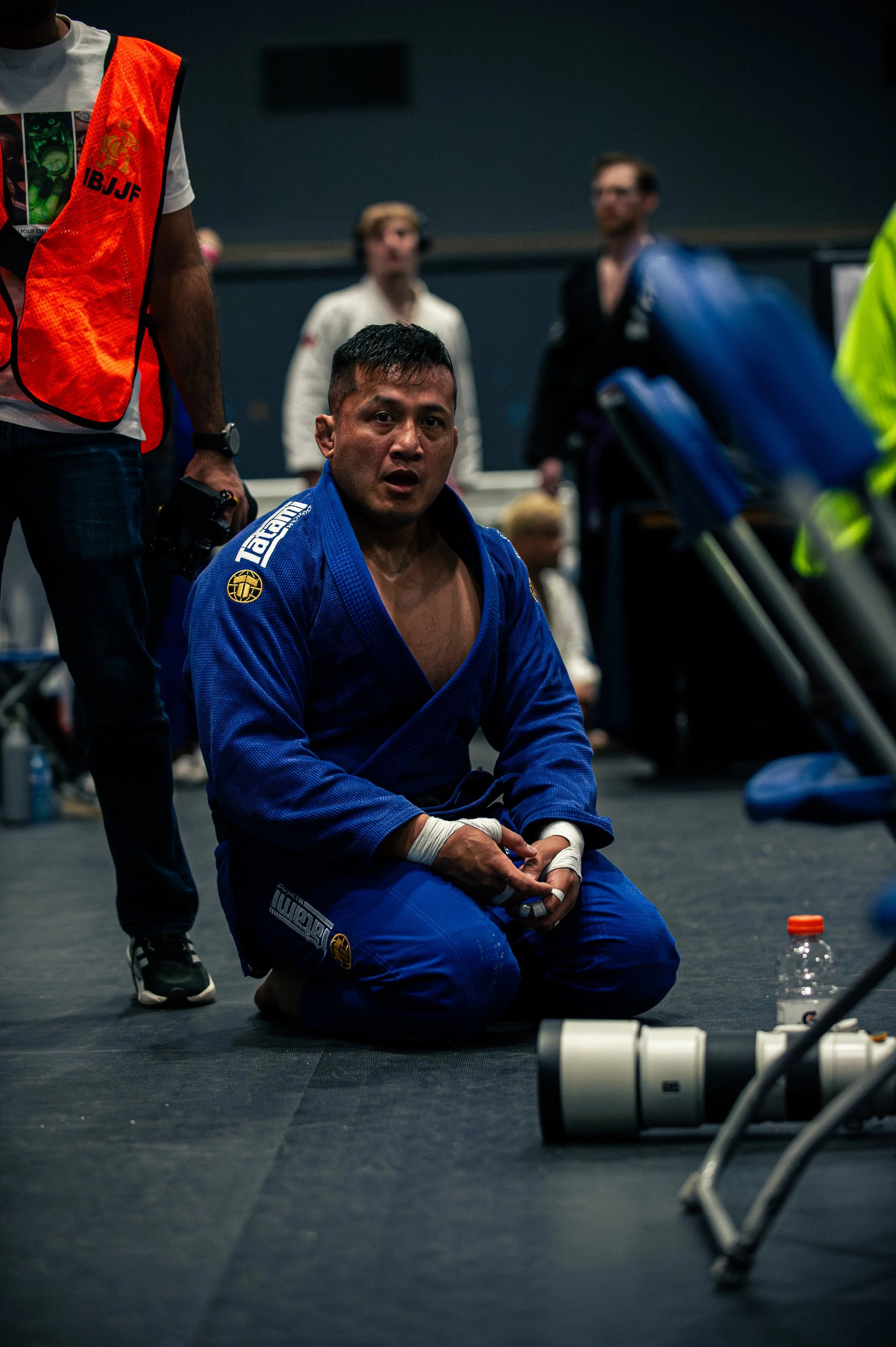 Male Brazilian Jiu-Jitsu competitor sitting on the floor, wearing a blue gi, surrounded by various equipment and other individuals in a training or competition setting.