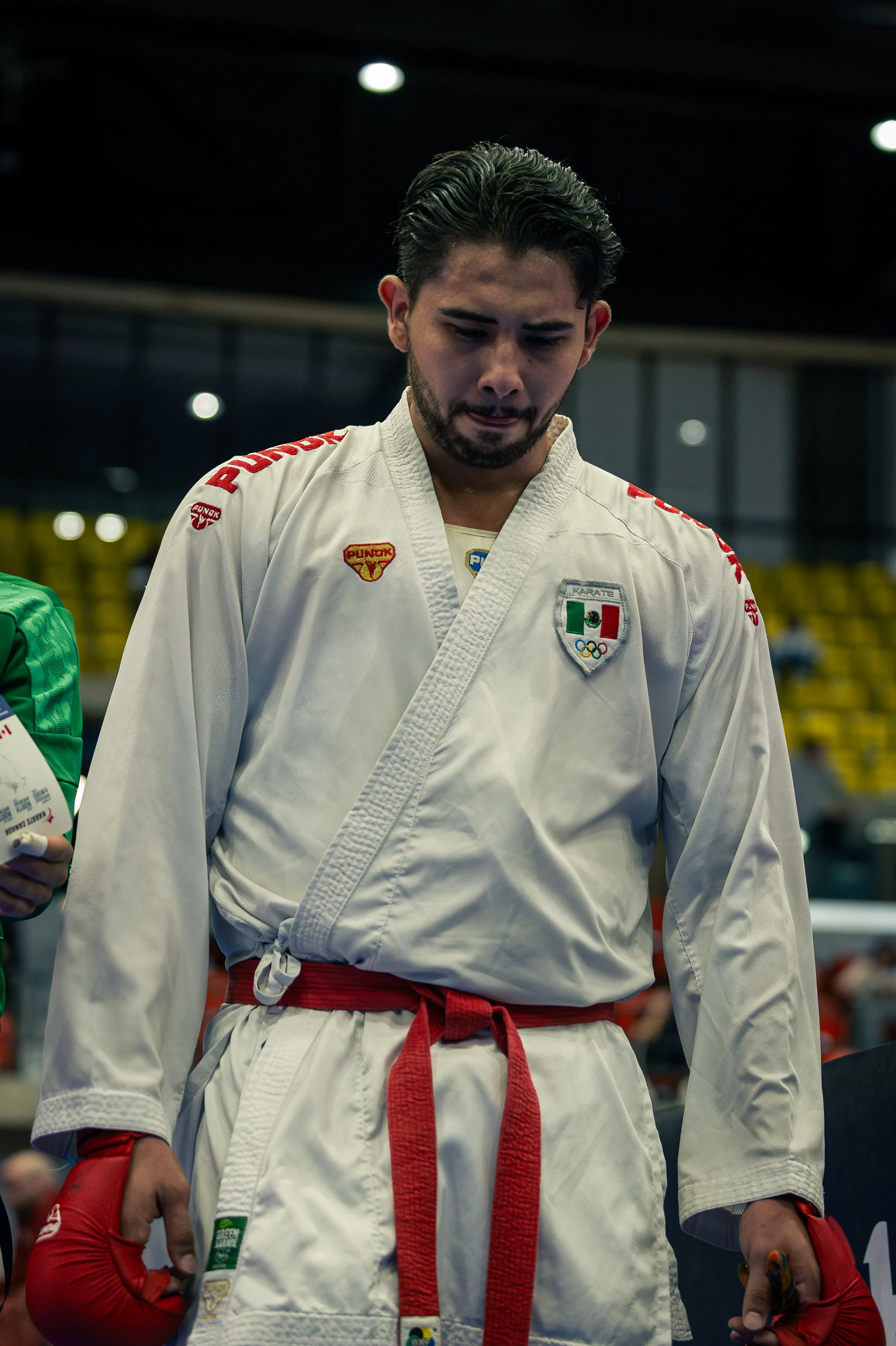 A male martial artist wearing a white karate gi with patches and a red belt, standing in an indoor venue, with a serious expression on his face. from Mexico