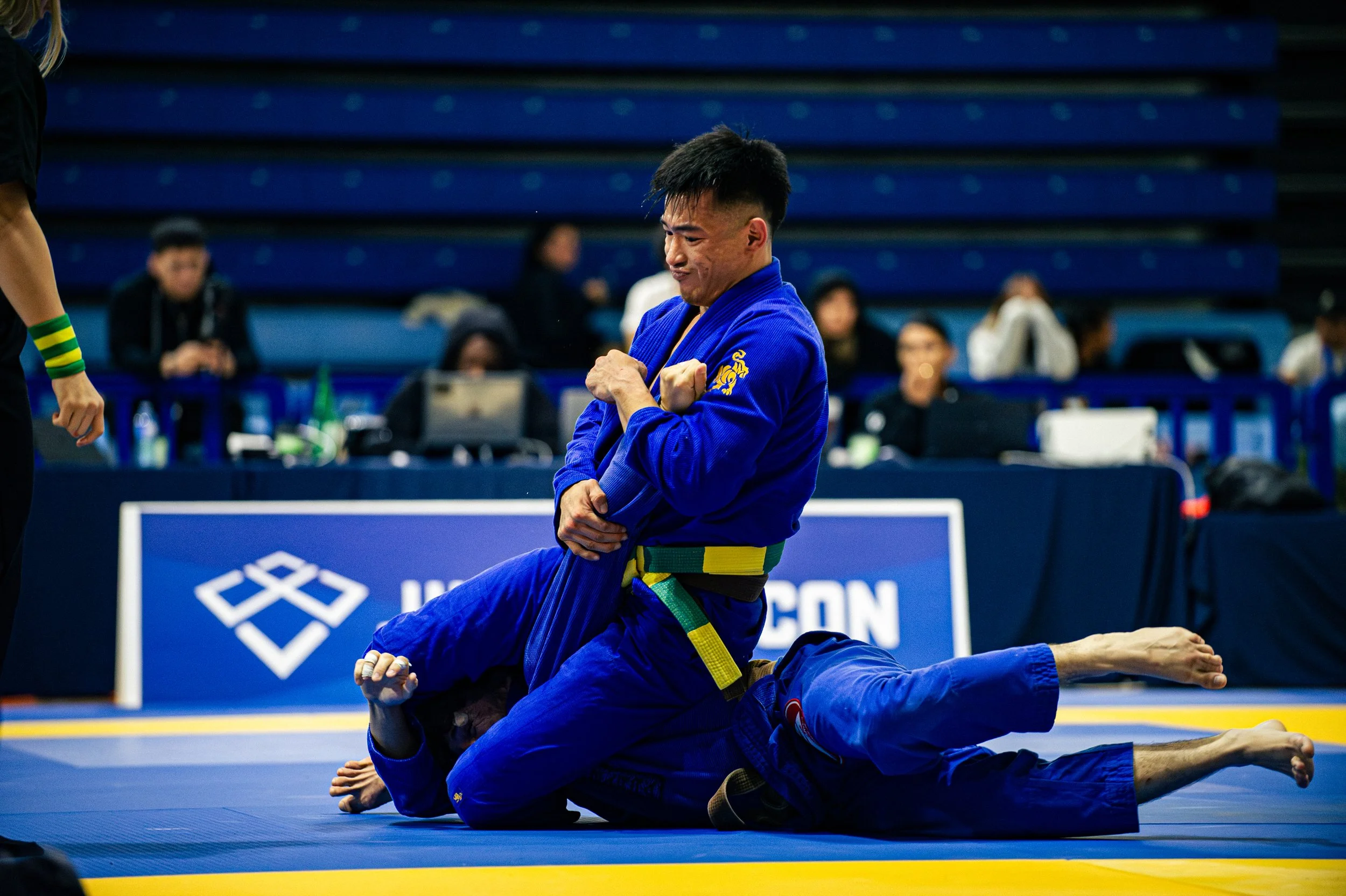 A male martial artist in a blue gi with a yellow belt demonstrates a submission hold during a competition, surrounded by a blue martial arts mat and spectators in the background.