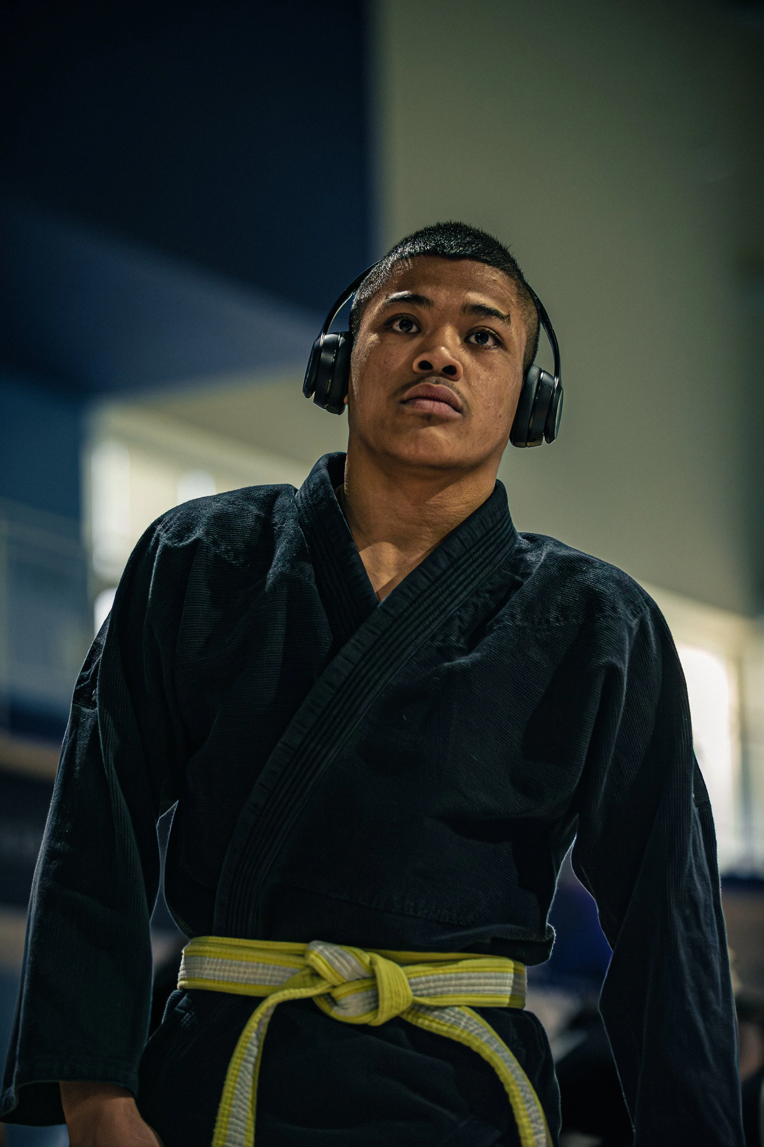 A young male martial artist wearing a black gi with a yellow belt, and headphones listening intently, appears focused.