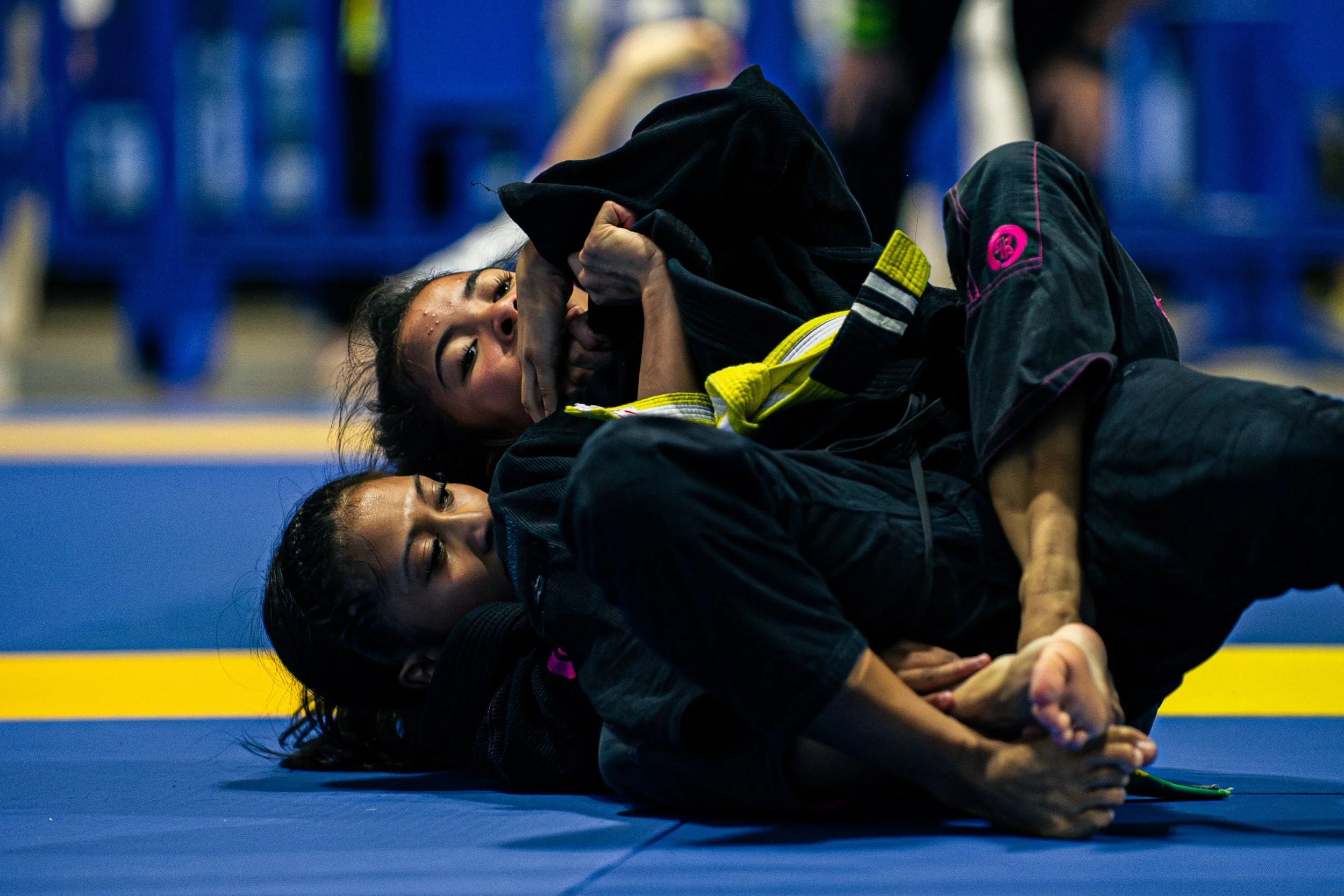 Two women practicing Brazilian Jiu-Jitsu on a blue mat, grappling and holding each other, with a blurred background of a gym or martial arts training facility.