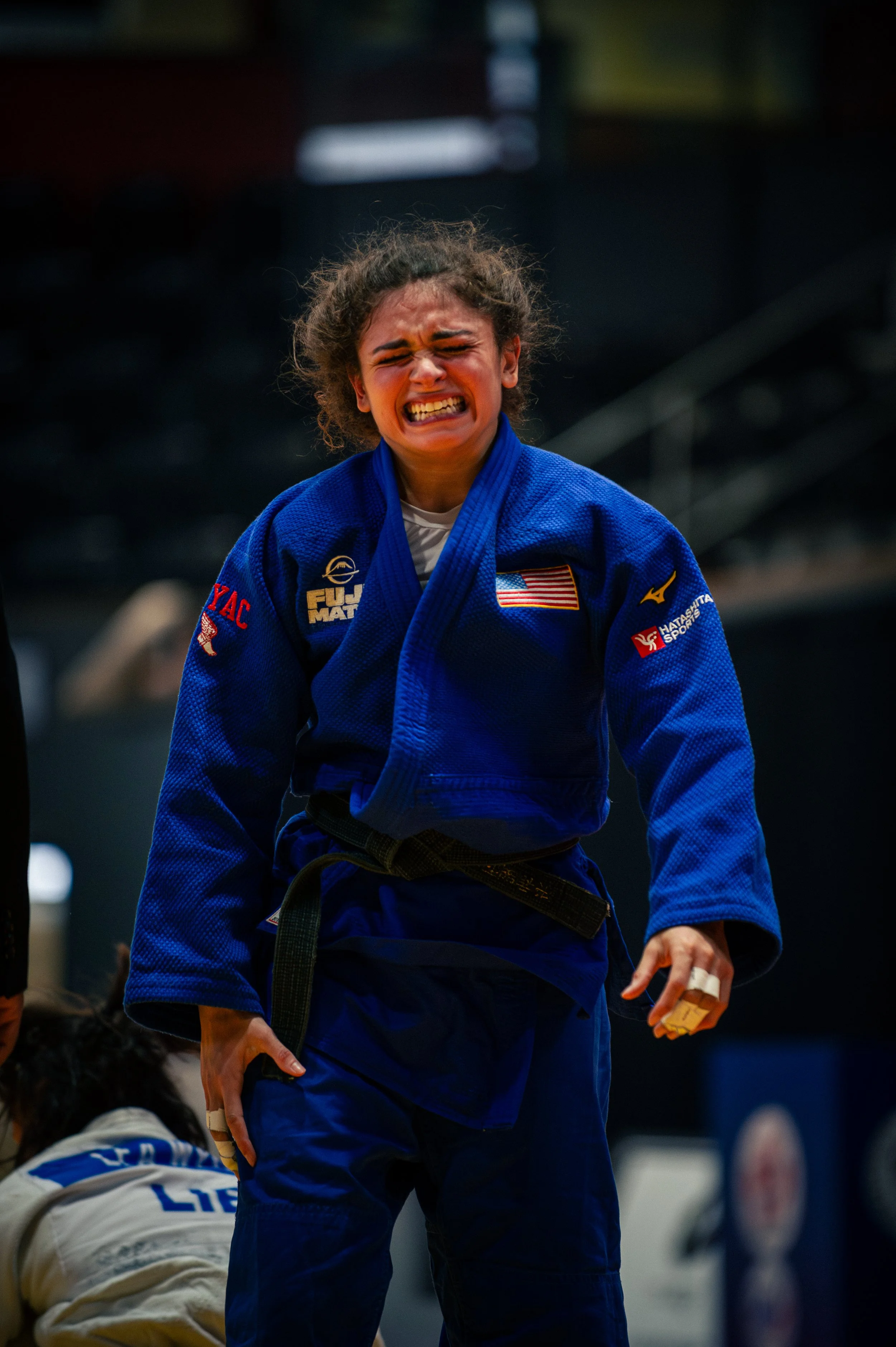 A female judo athlete in a blue gi with patches, struggling during a match, with a person in a white gi on the ground in the background.