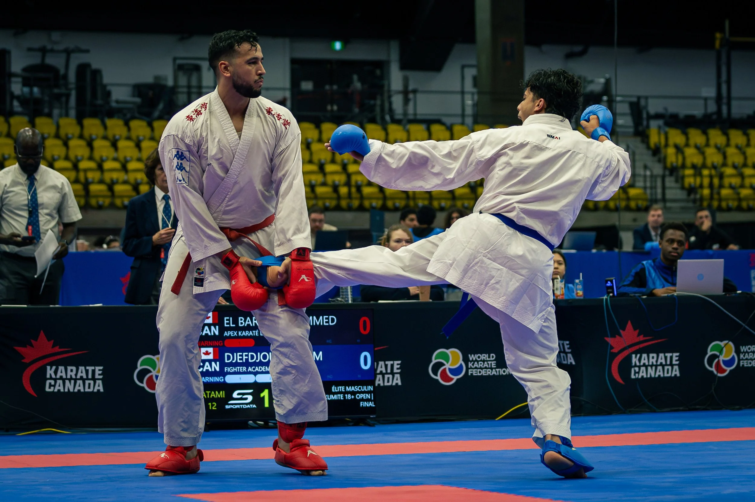 Two martial artists wearing traditional karate gi and gloves sparring in a competition, with a referee and an audience in the background at a karate tournament.