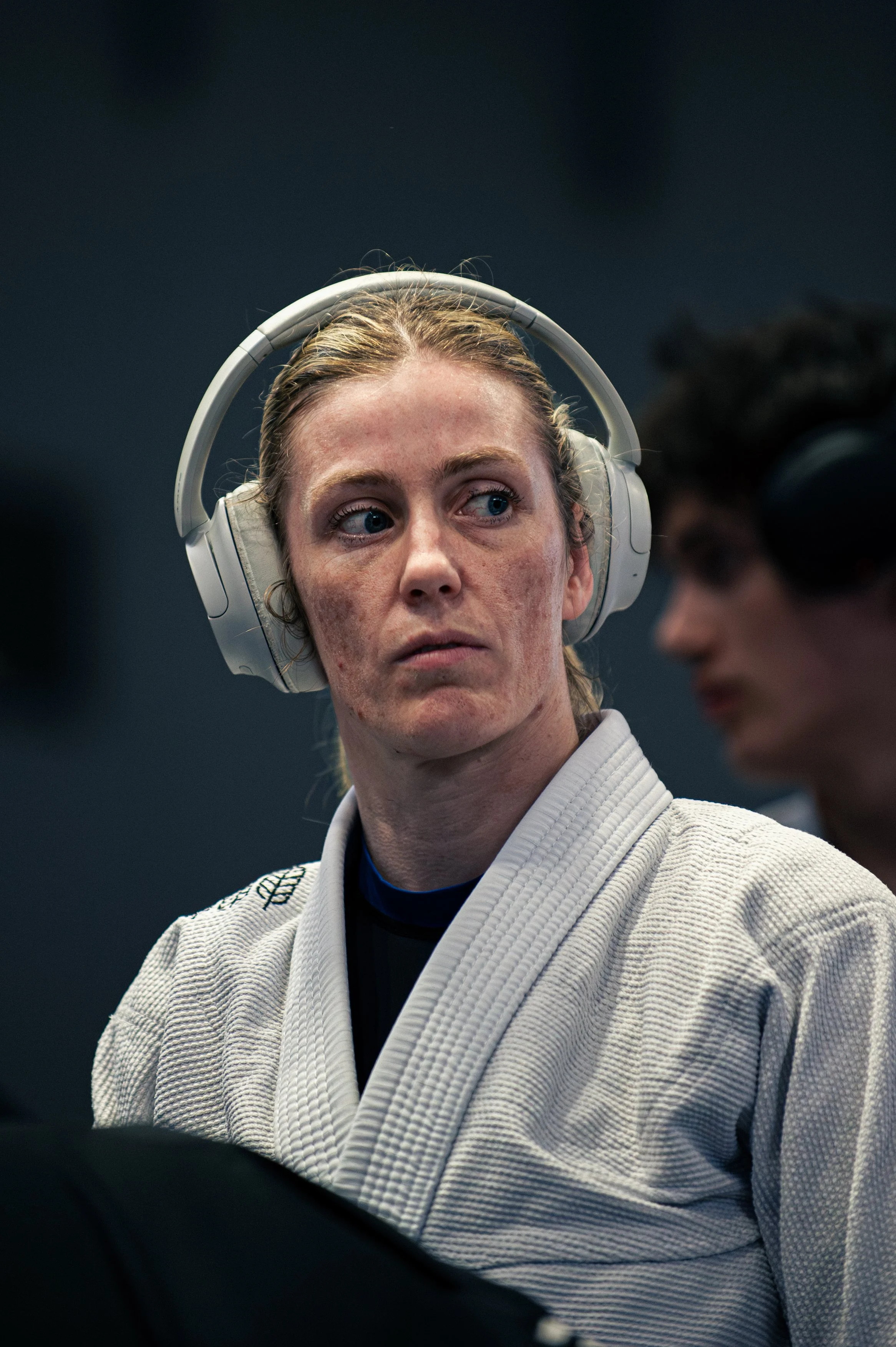 Close-up of a woman wearing a white jbjj gi and a white headset, looking to her left with a serious expression.