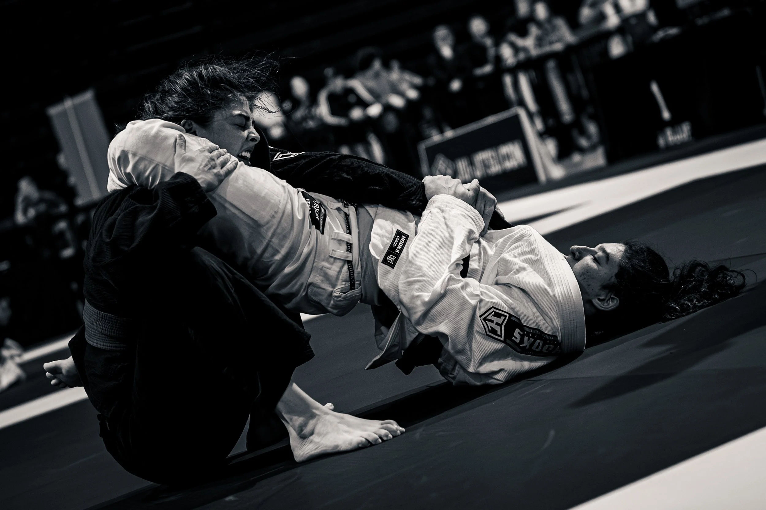 Two women practicing Brazilian Jiu-Jitsu on mats in a gym. One woman, wearing a white gi with patches, is on her back, gripping the collar of her opponent, who is in a black gi. The black gi woman is leaning over her opponent in a defensive posture.