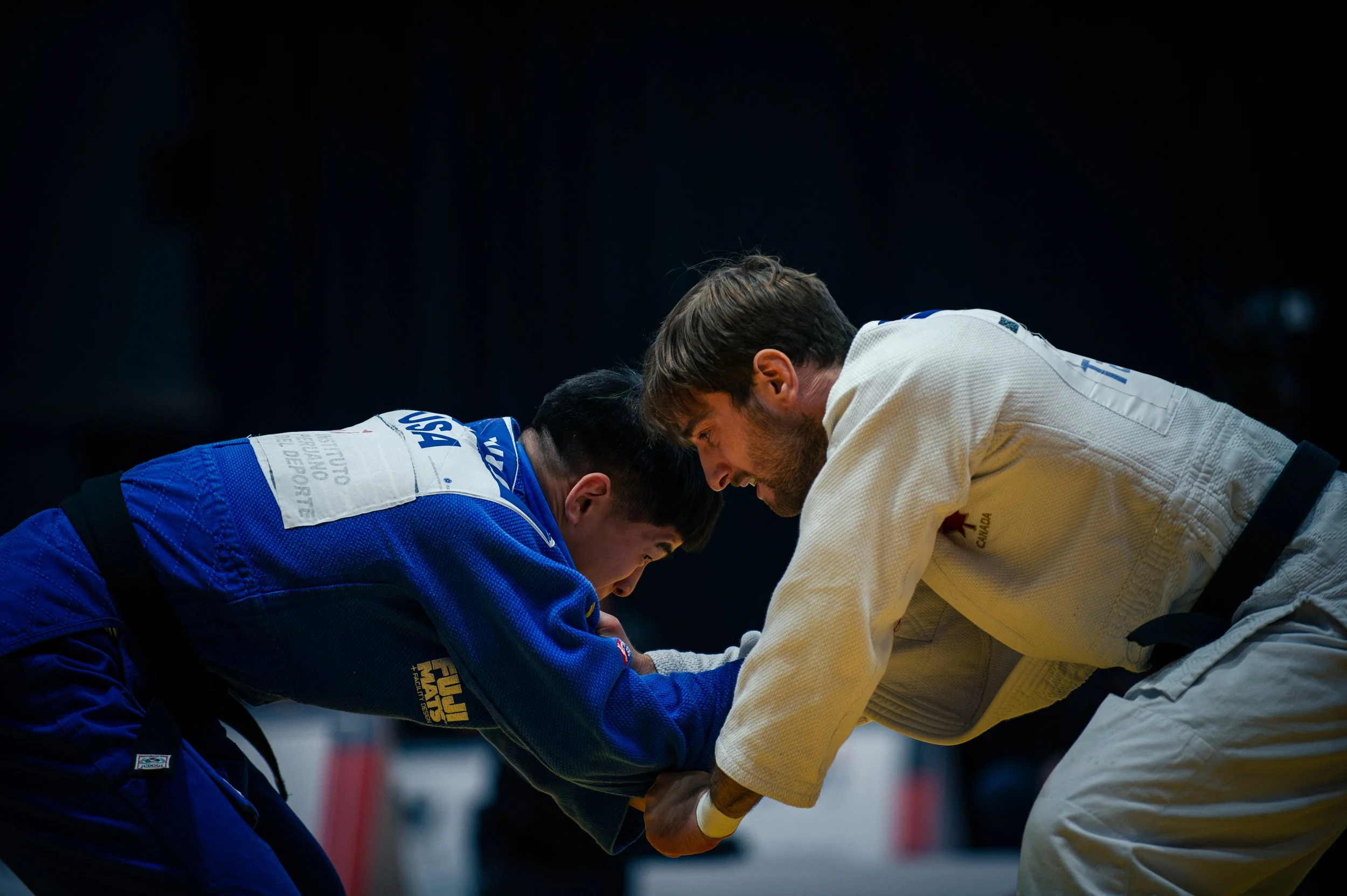 Two judo competitors, one in a blue gi and the other in a white gi, bent forward and holding each other's arms during a match. IFJ Judo Canada