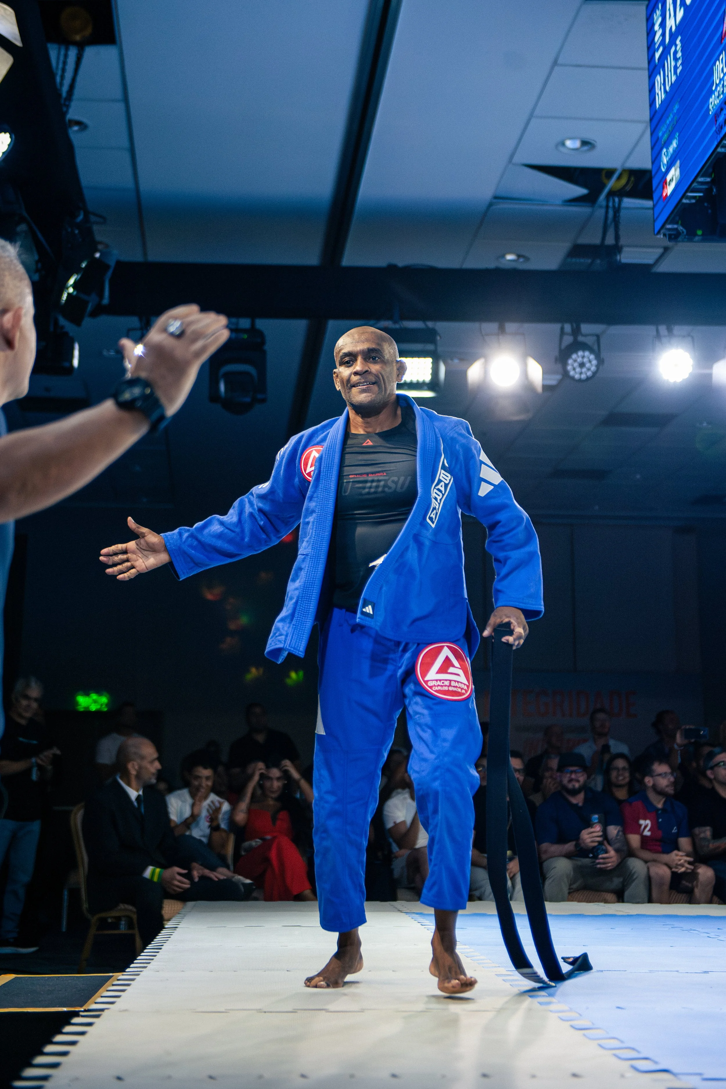 A male Brazilian Jiu-Jitsu competitor in a blue gi, standing on a mat during a match with his arms outstretched. He is in front of an audience and bright overhead lights.