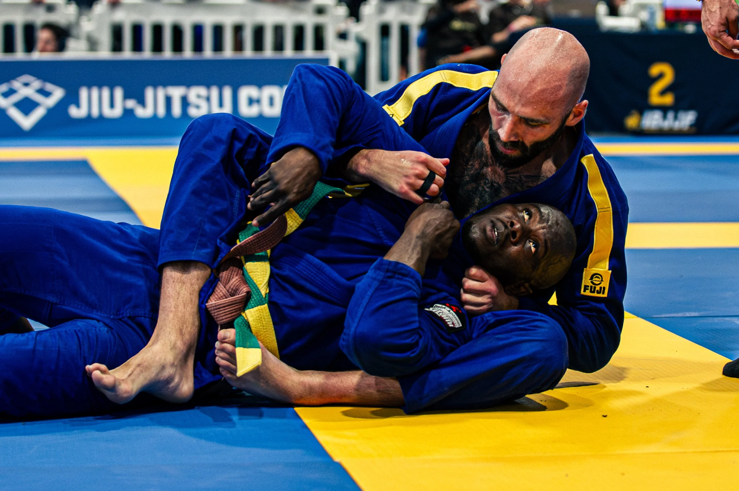 Two male Brazilian Jiu-Jitsu competitors on a yellow and blue mat during a match, with one athlete on top and the other on the ground, both wearing blue gis. The athlete on top has a bald head and a beard. The background shows a banner with the text 