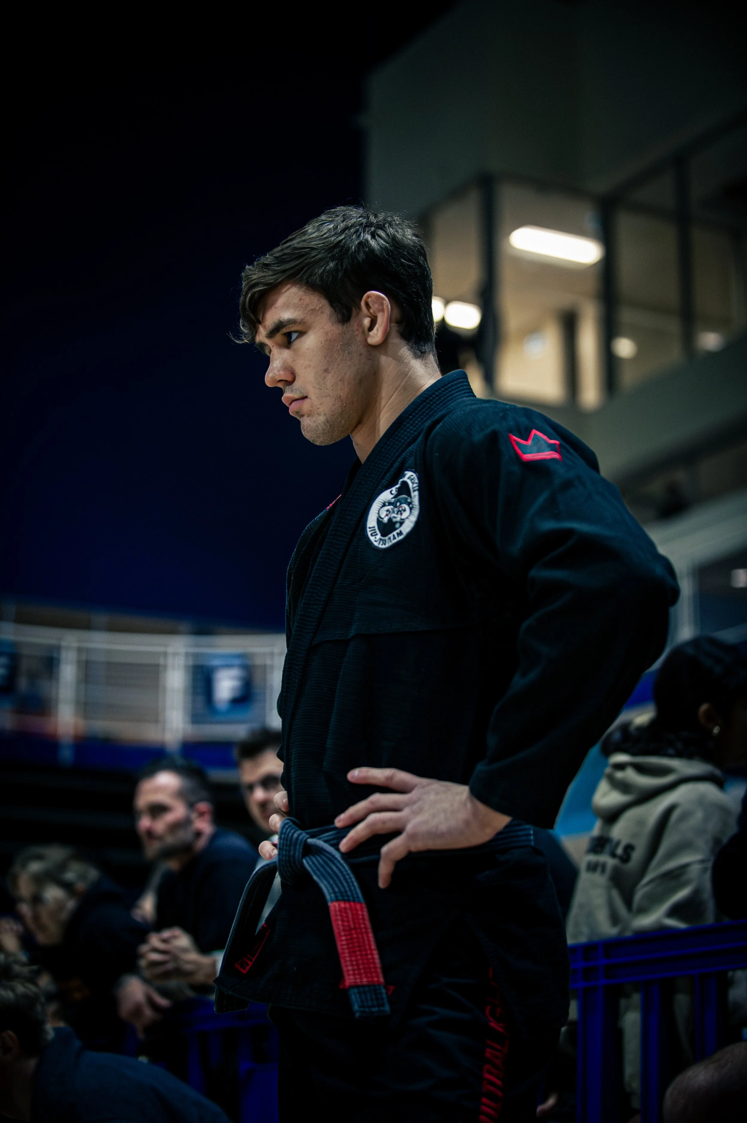 A young male martial artist in a black gi with patches and a black belt, standing attentively with hands on hips, in an indoor gym with spectators in the background. IBJJF Toronto