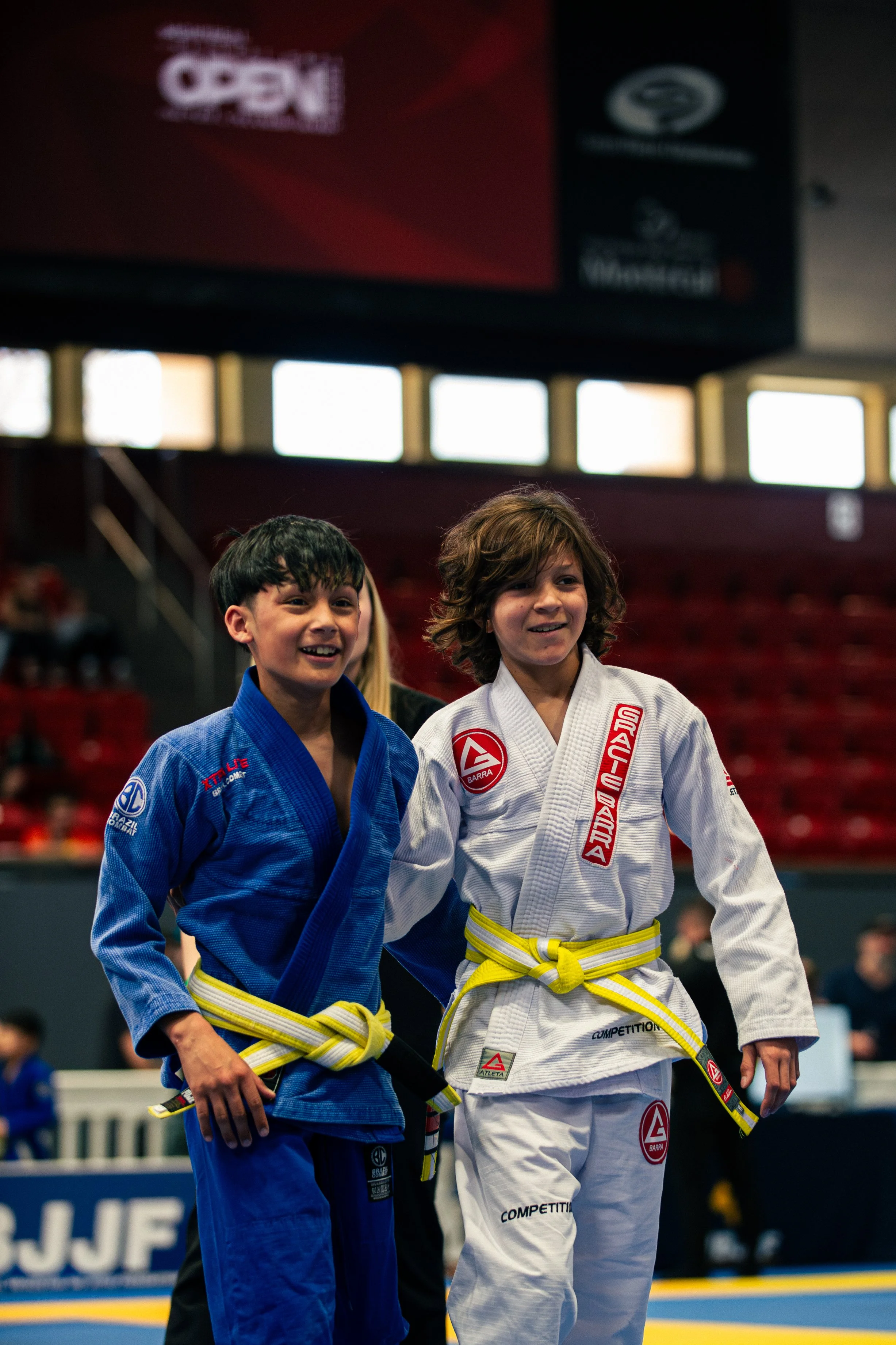 Two young boys in Brazilian Jiu-Jitsu uniforms smiling and posing together in a sports arena, one in a blue gi and the other in a white gi with a yellow belt.