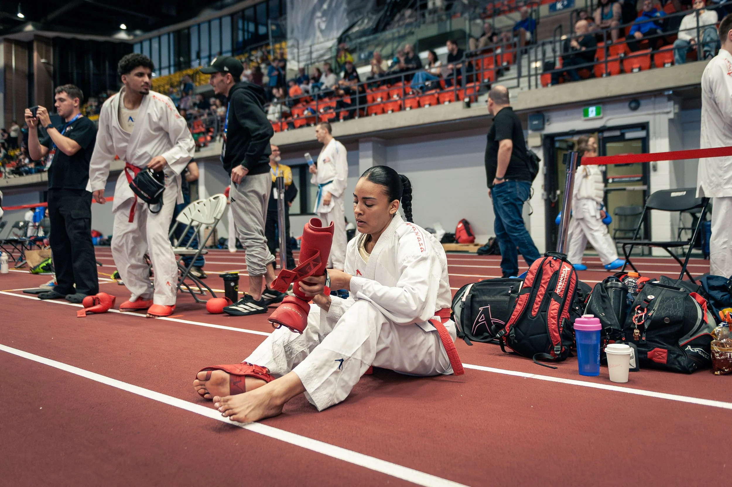 A female martial artist in a white gi with a red belt, sitting on the ground, holding red sparring gear, surrounded by equipment and bags indoors at a sports stadium with spectators in the background.