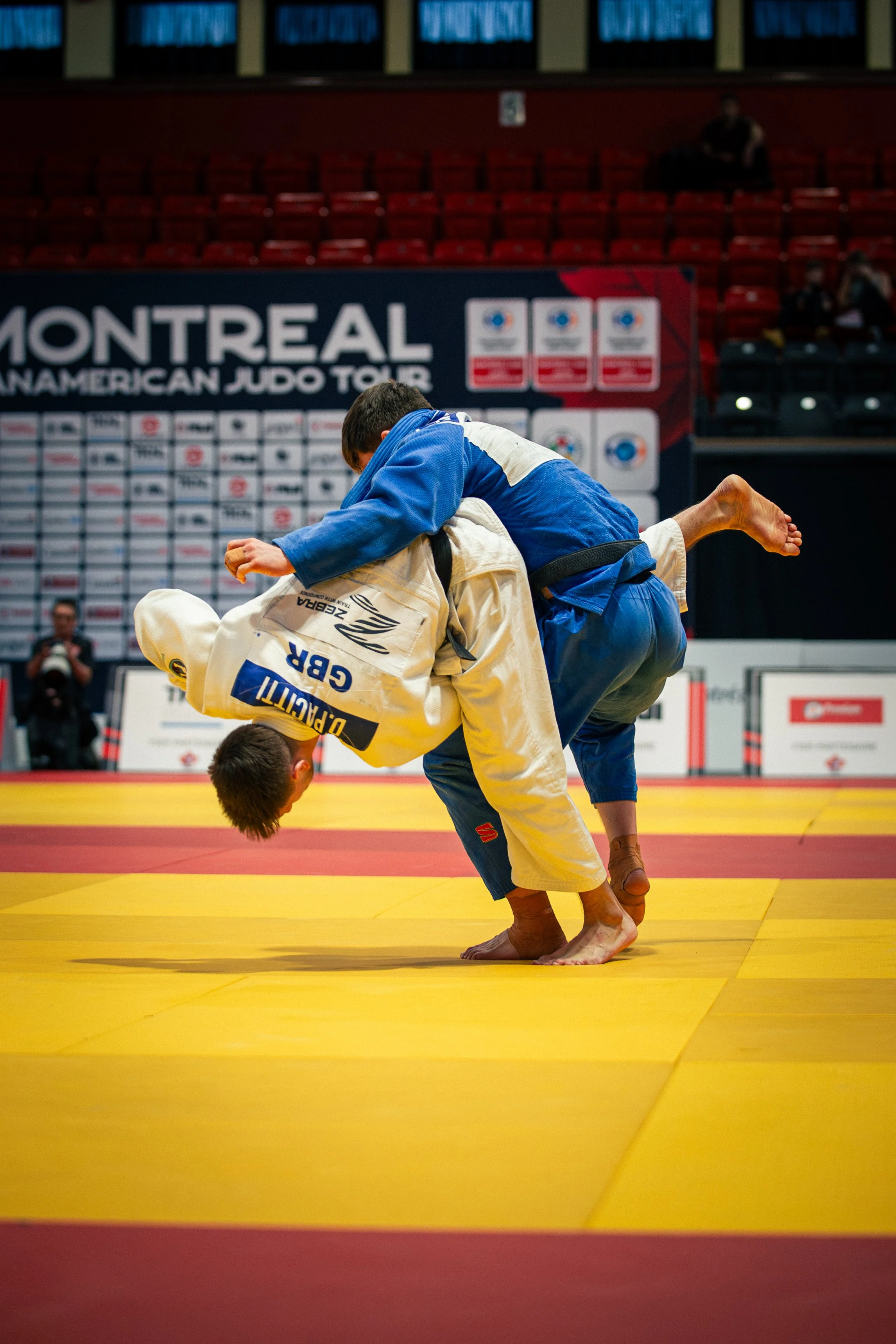 Two male judo competitors in blue and white gis engaged in a match on a yellow mat at a tournament with a Montreal Judo Tour banner in the background.