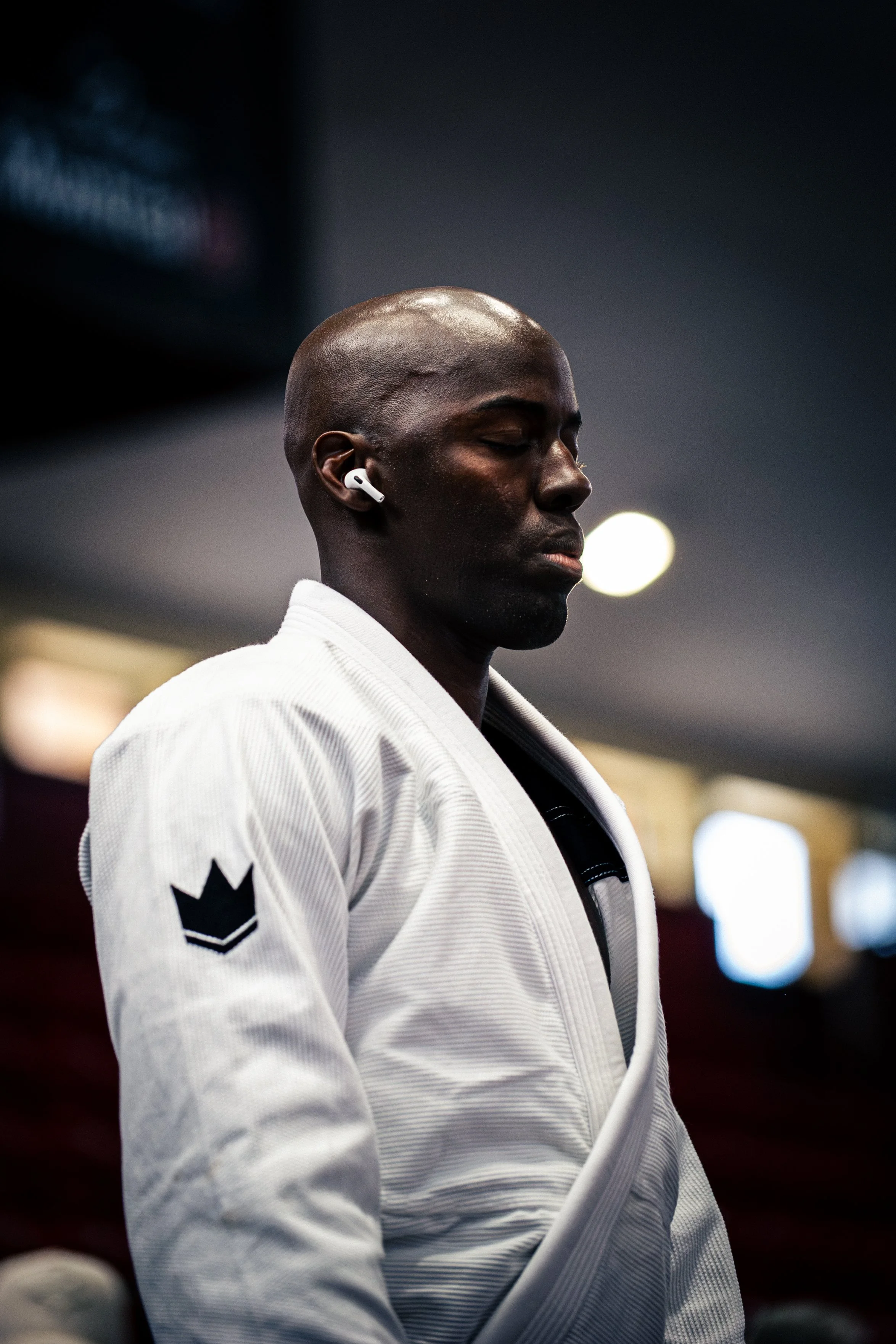 A person with dark skin and a shaved head wearing a white judo gi with a black star on the sleeve and an emblem on the chest, eyes closed, possibly meditating or resting, with a blurred background and bright lights.