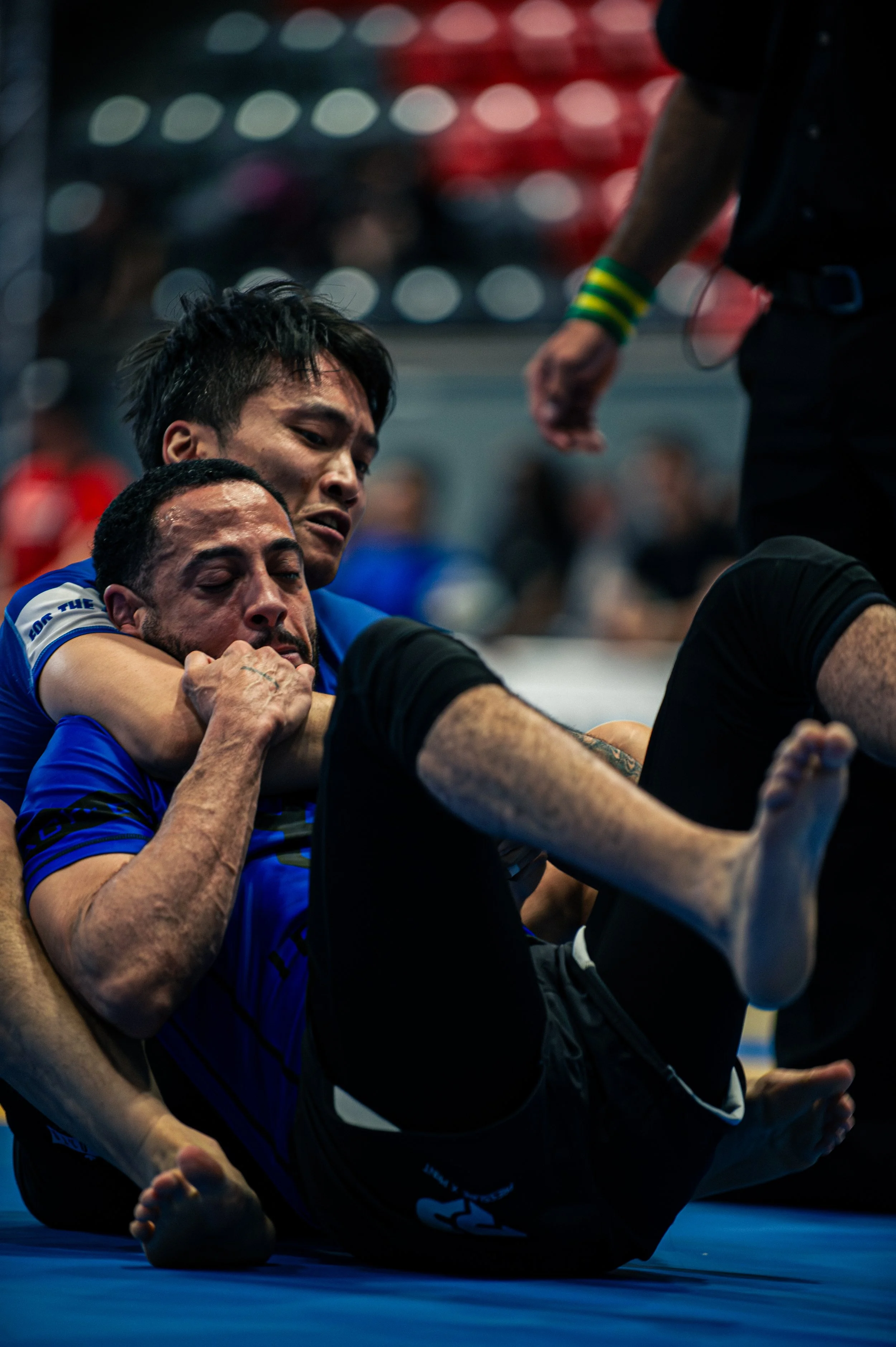 Two men engaged in a Brazilian Jiu-Jitsu match on a blue mat, one applying a chokehold on the other, with a referee watching attentively.