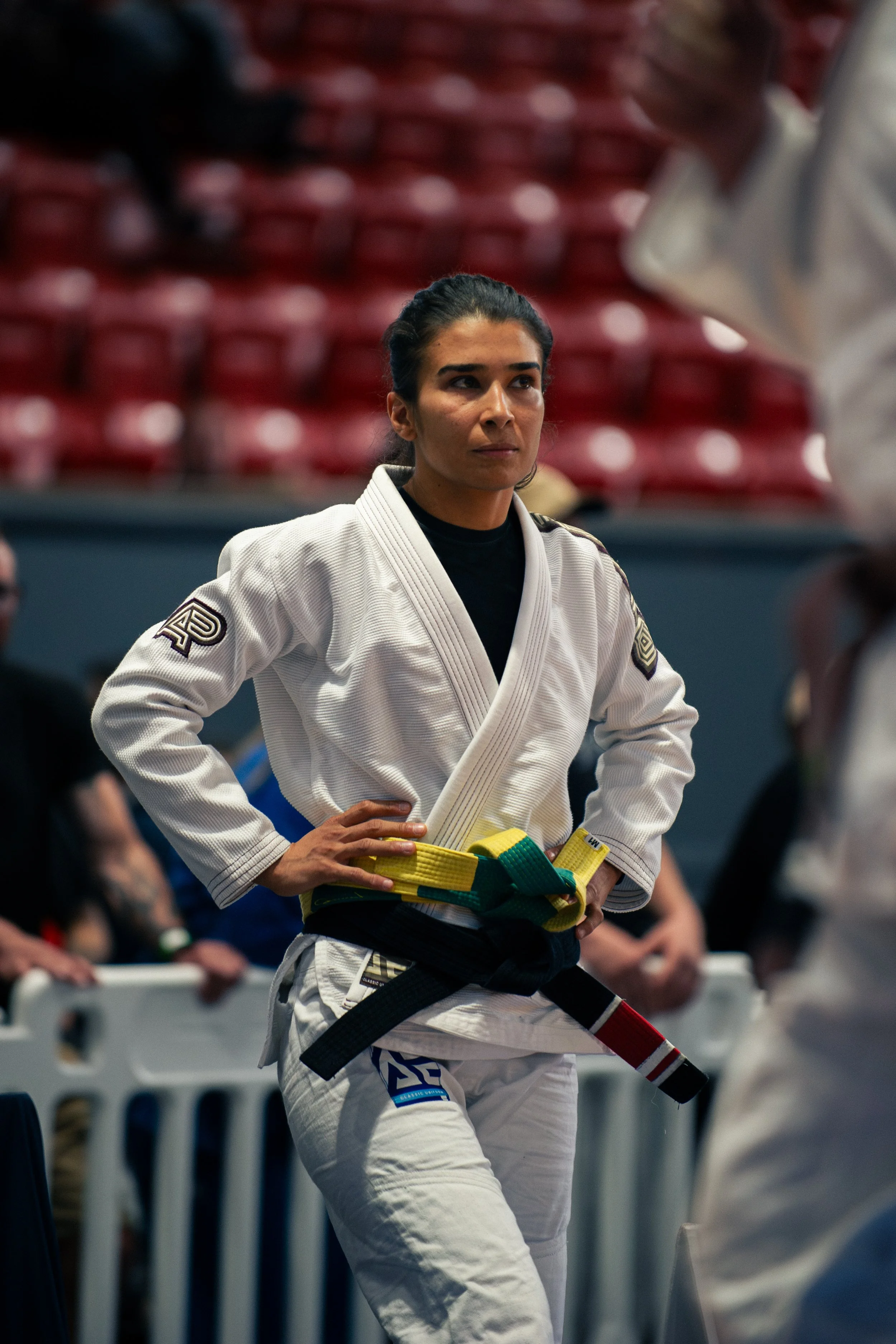 Female martial artist in a white gi with a black belt, standing with hands on hips in a gym or dojo with red seating in the background.