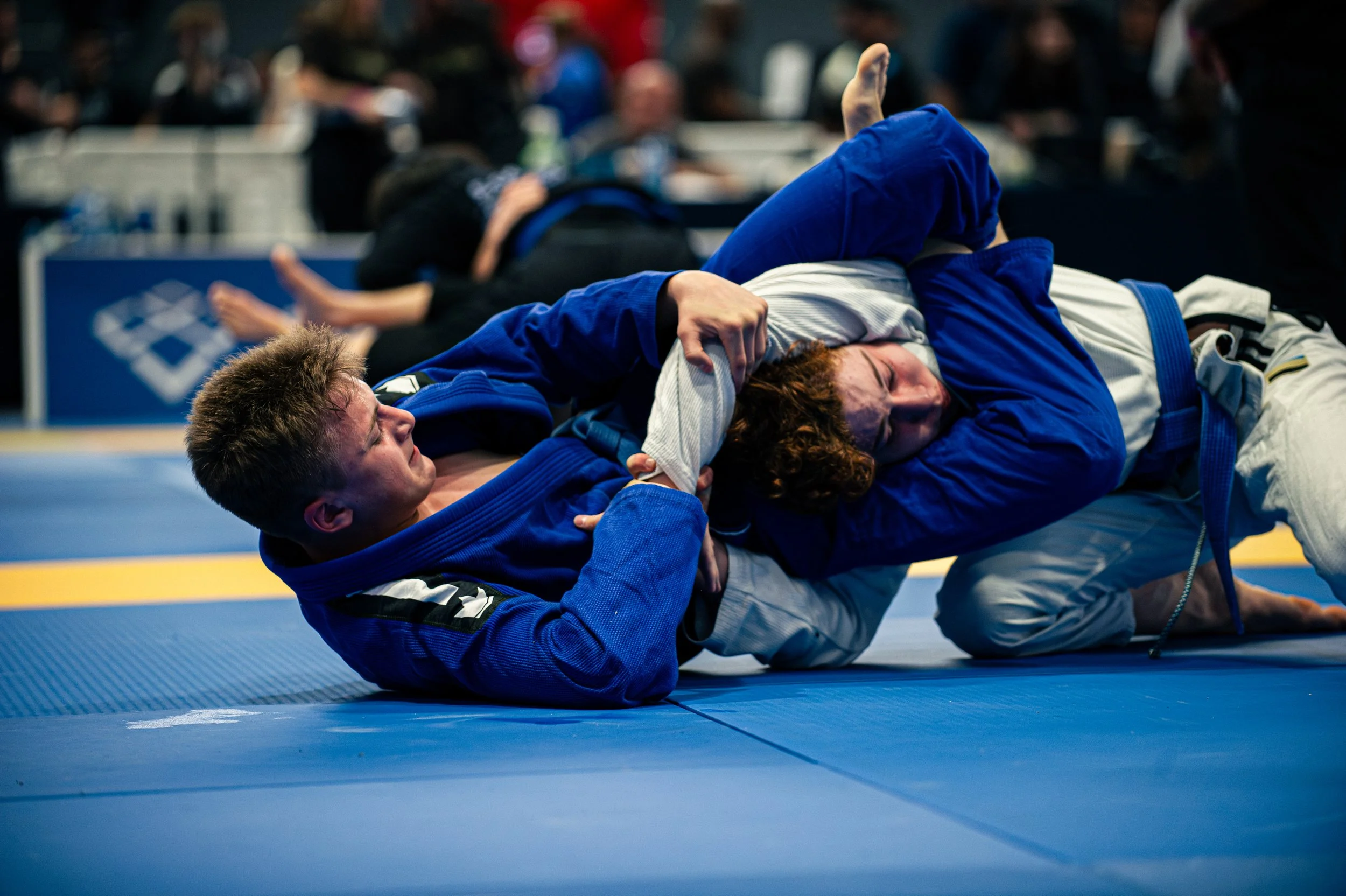 Two Brazilian Jiu-Jitsu practitioners in blue gis engaged in a grappling match on a blue and yellow mat. One person is on top, attempting a submission, while the other is on the ground trying to defend. The background shows a blurred audience and com