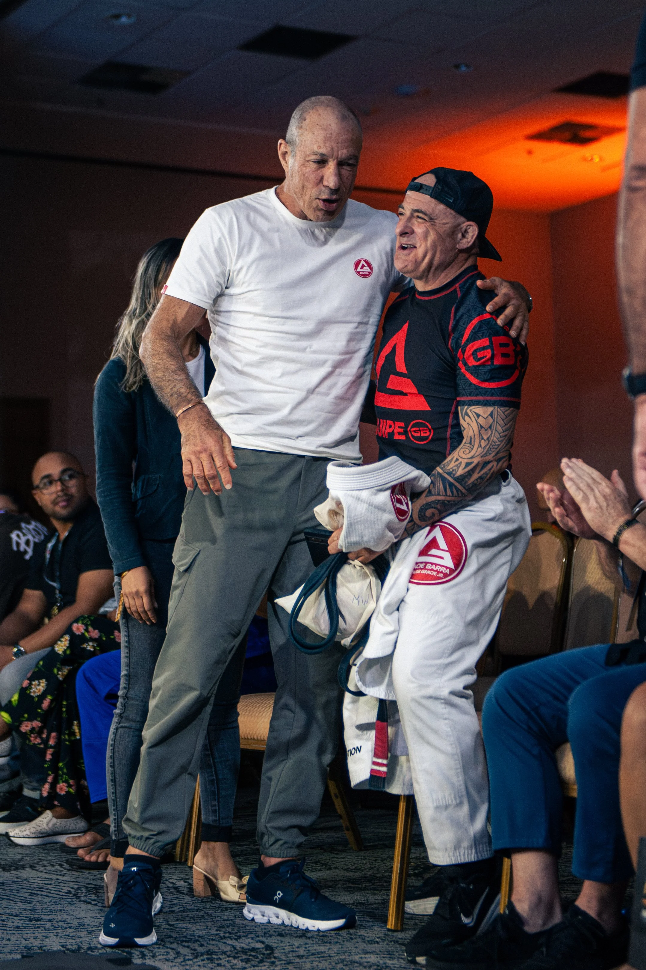  one in a white t-shirt and gray pants, and another in a black and red sports uniform, are standing closely together, engaging in conversation. The man in sports attire is holding a white gi with a logo, and they are surrounded by seated people in a 