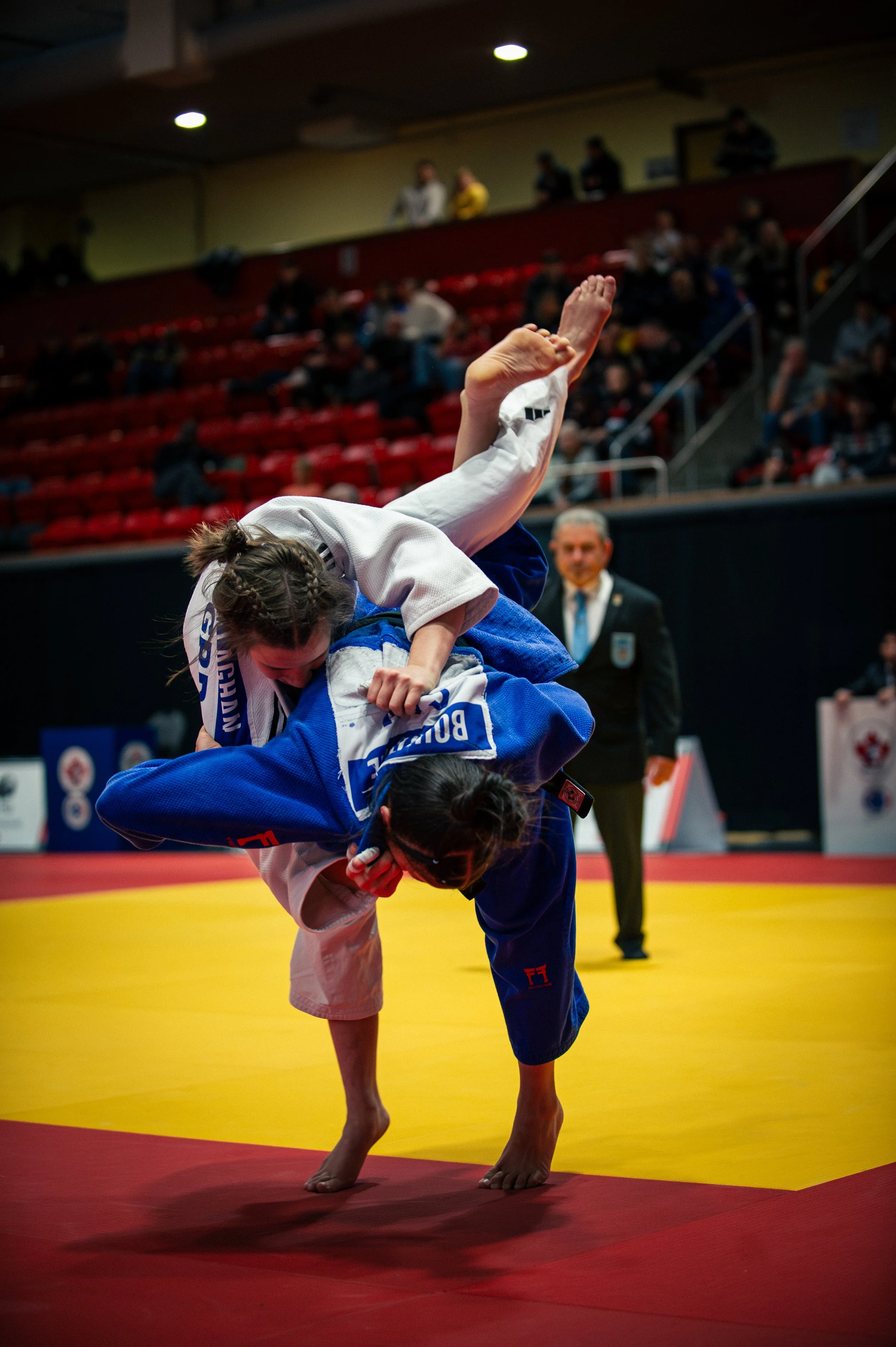 Two female judo competitors in action during a match on a yellow mat, with a referee in the background and spectators watching from the stands.