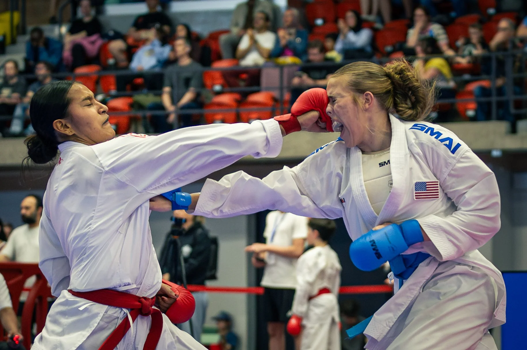 Two female martial artists competing in a karate competition open international de Montreal, punching each other in the face, with an audience watching in the background.