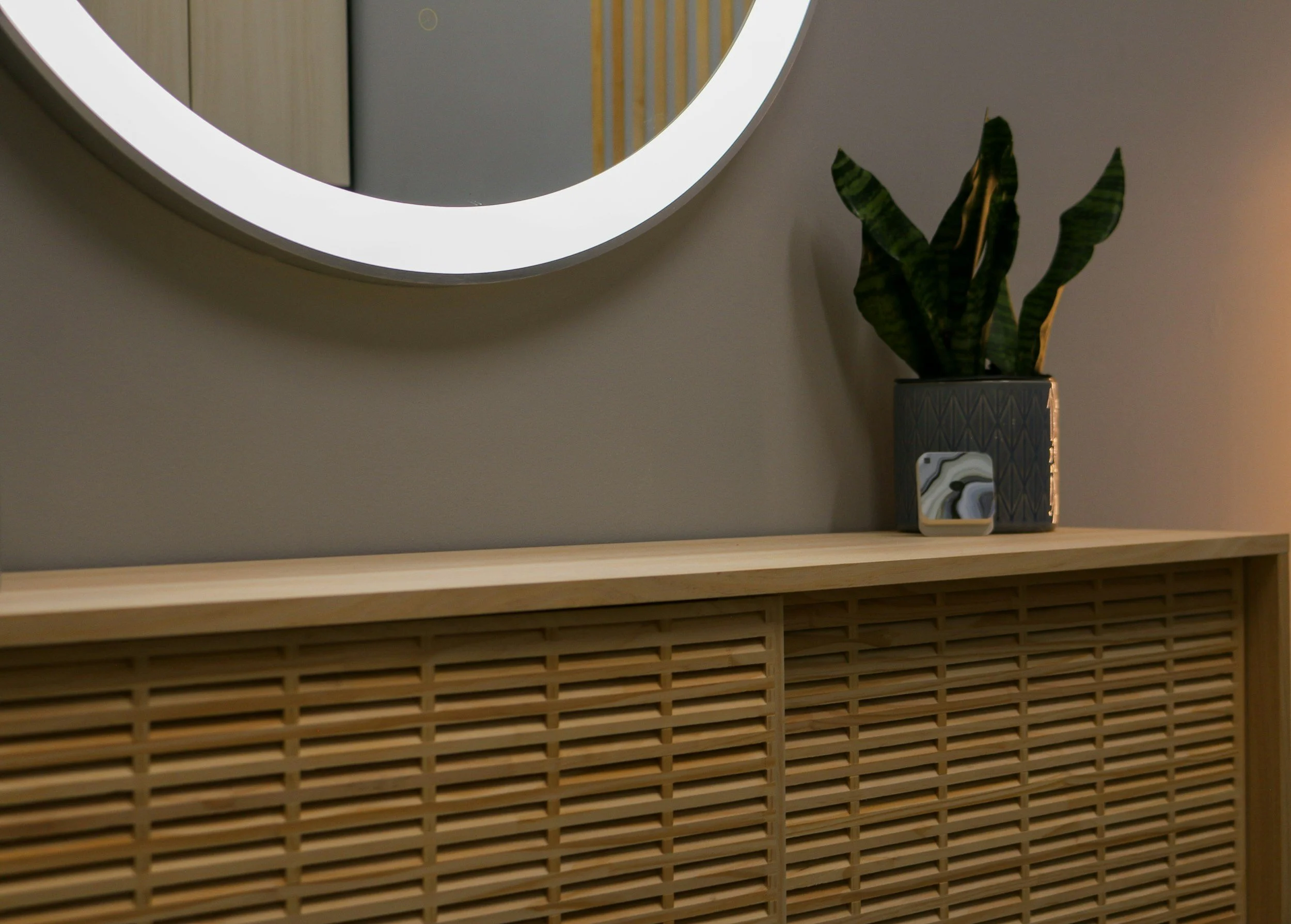 A round LED mirror reflecting a part of a wall with a door and yellow-striped design, a potted plant with dark green patterned leaves, and a smartphone placed on a wooden surface in front of the plant.