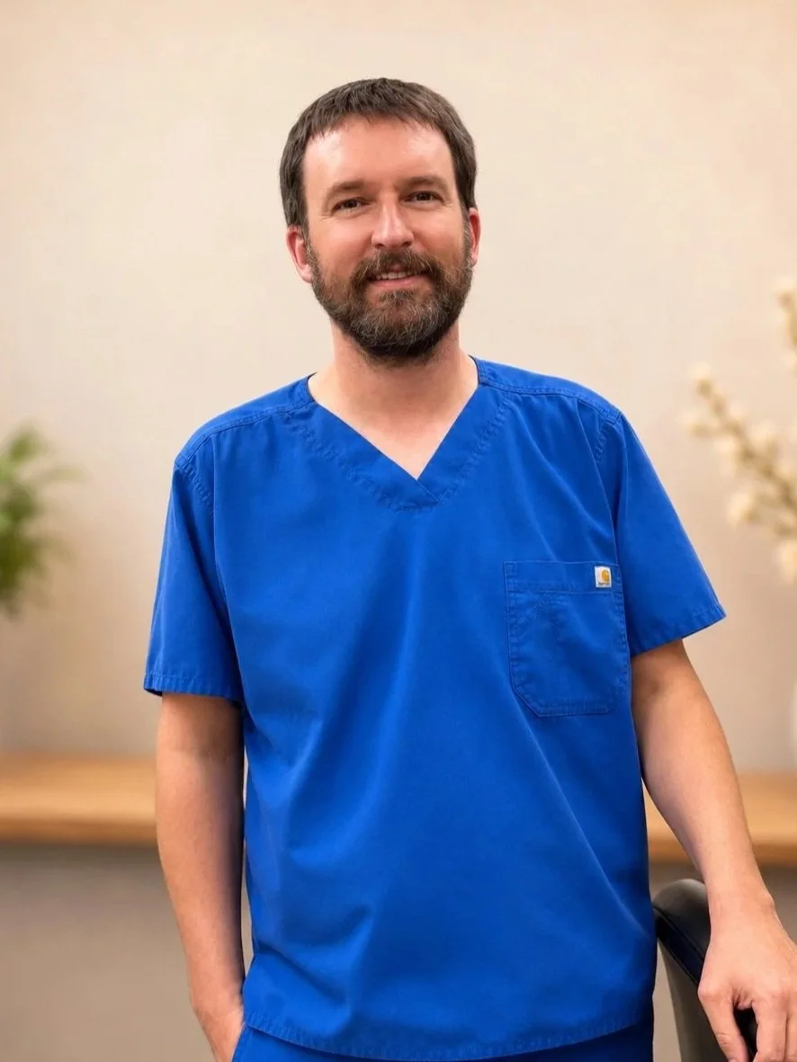 A man with a beard wearing blue medical scrubs standing indoors, smiling at the camera.
