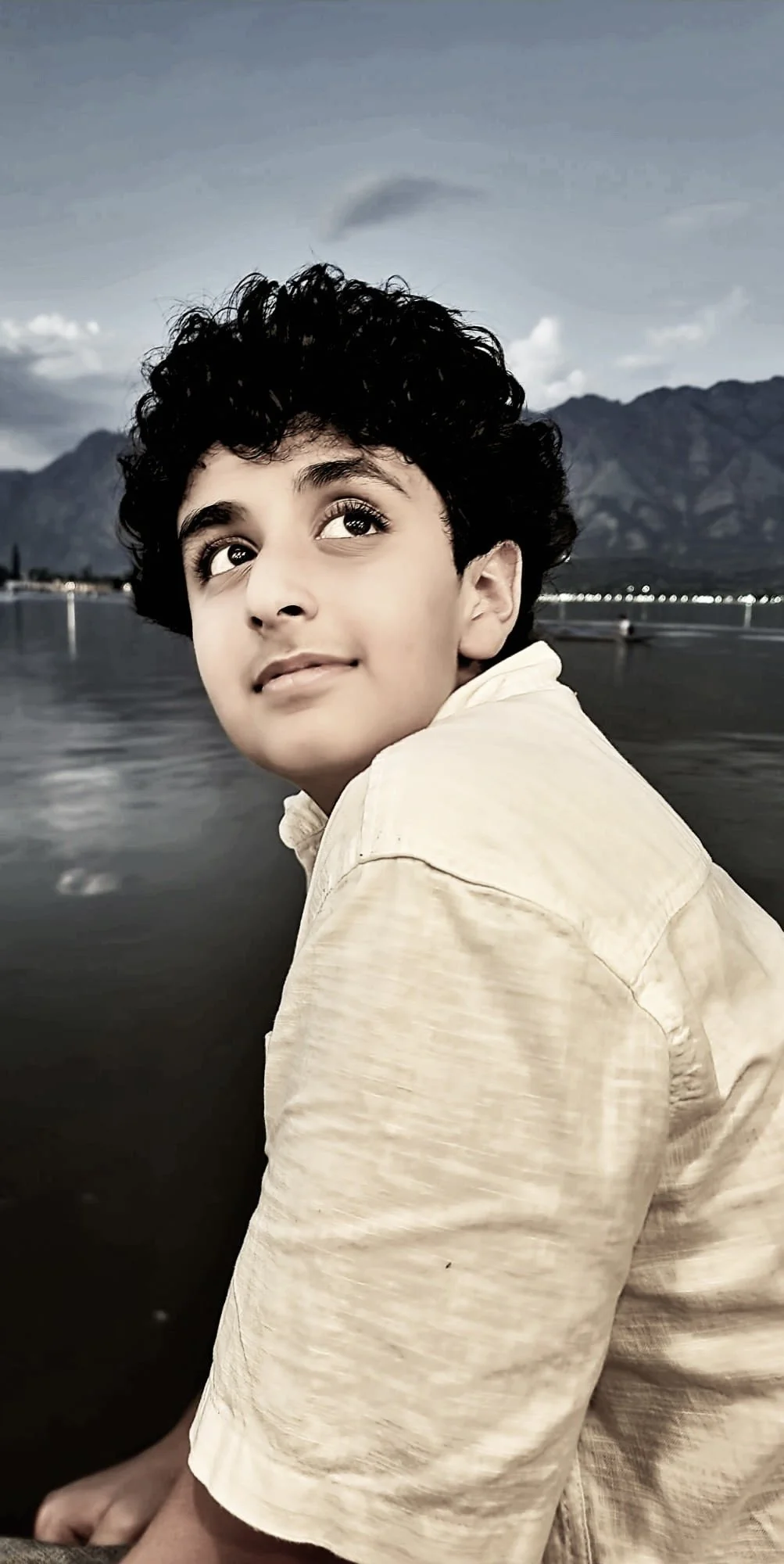 A young boy with curly black hair and light skin sitting outdoors near a body of water, with mountains and a cloudy sky in the background.