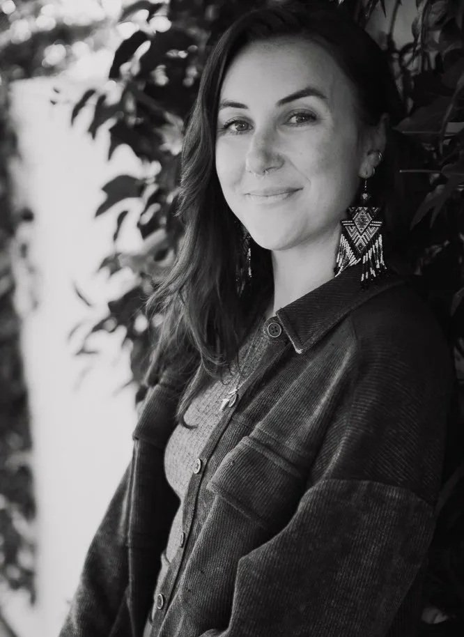 A woman with long hair smiling at the camera, wearing statement earrings and a denim jacket, standing beside leafy plants.