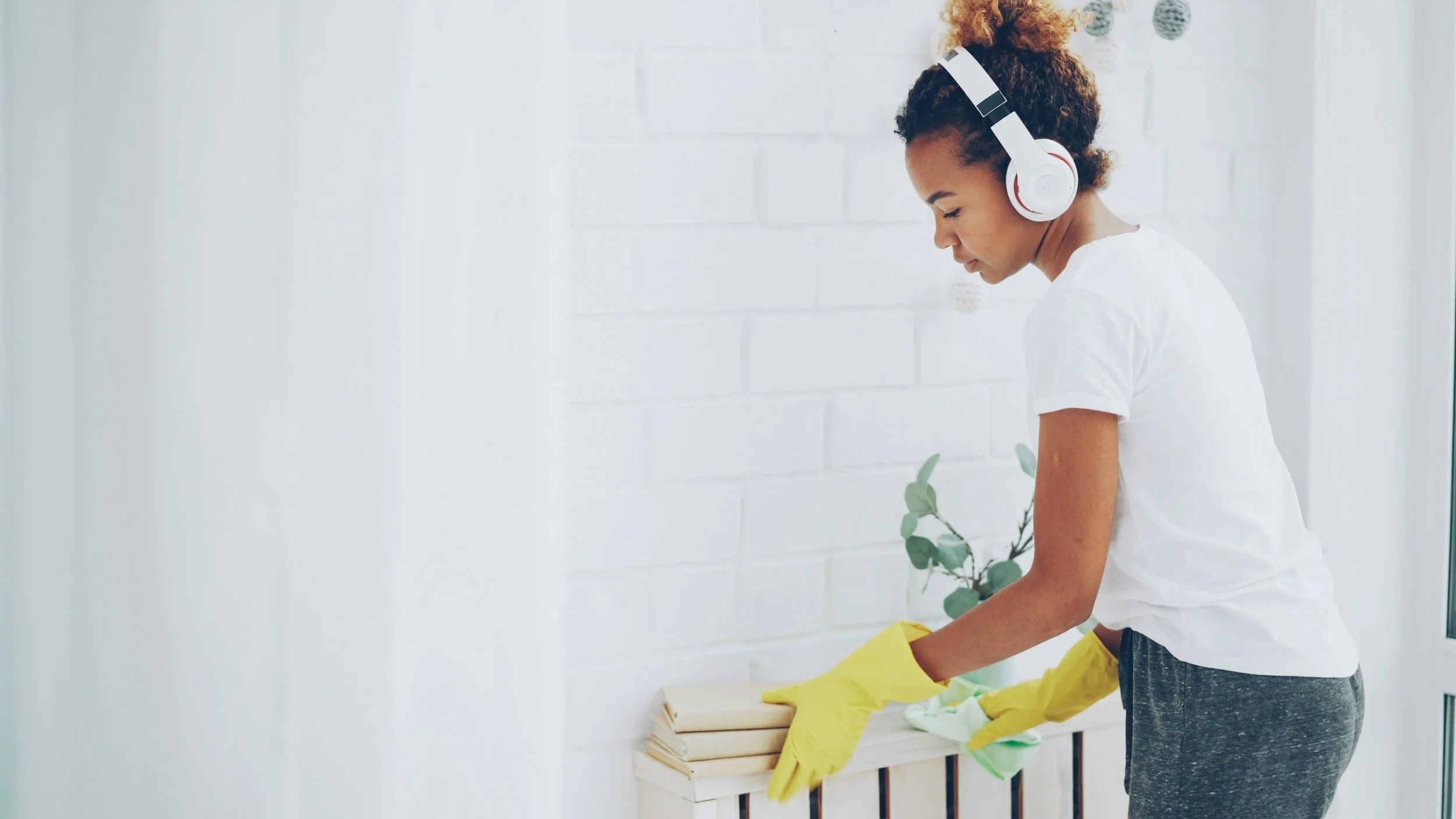 A woman wearing headphones and a white t-shirt is cleaning or gardening, holding yellow gloves and a green cloth, with a small green plant on a white wooden stand in front of her, against a white brick wall.