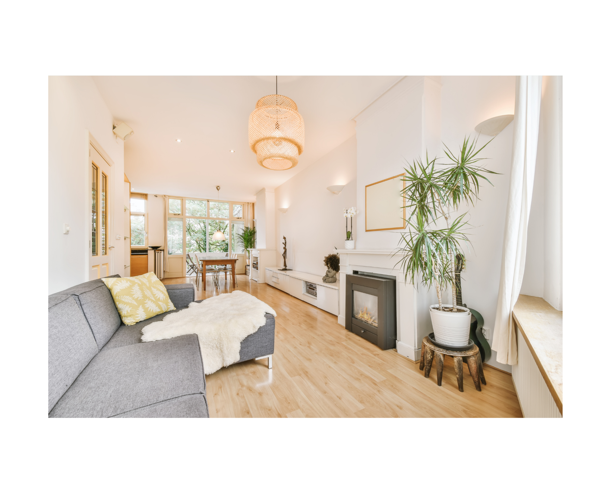 Bright modern living room with gray sofa, white walls, wooden flooring, large window, potted plant, fireplace, and woven pendant light.