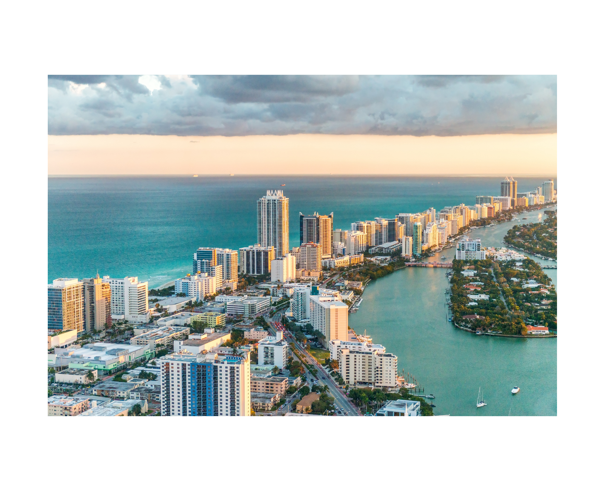 Aerial view of a coastal city with high-rise buildings, a waterway with boats, and a cloudy sky at sunset.