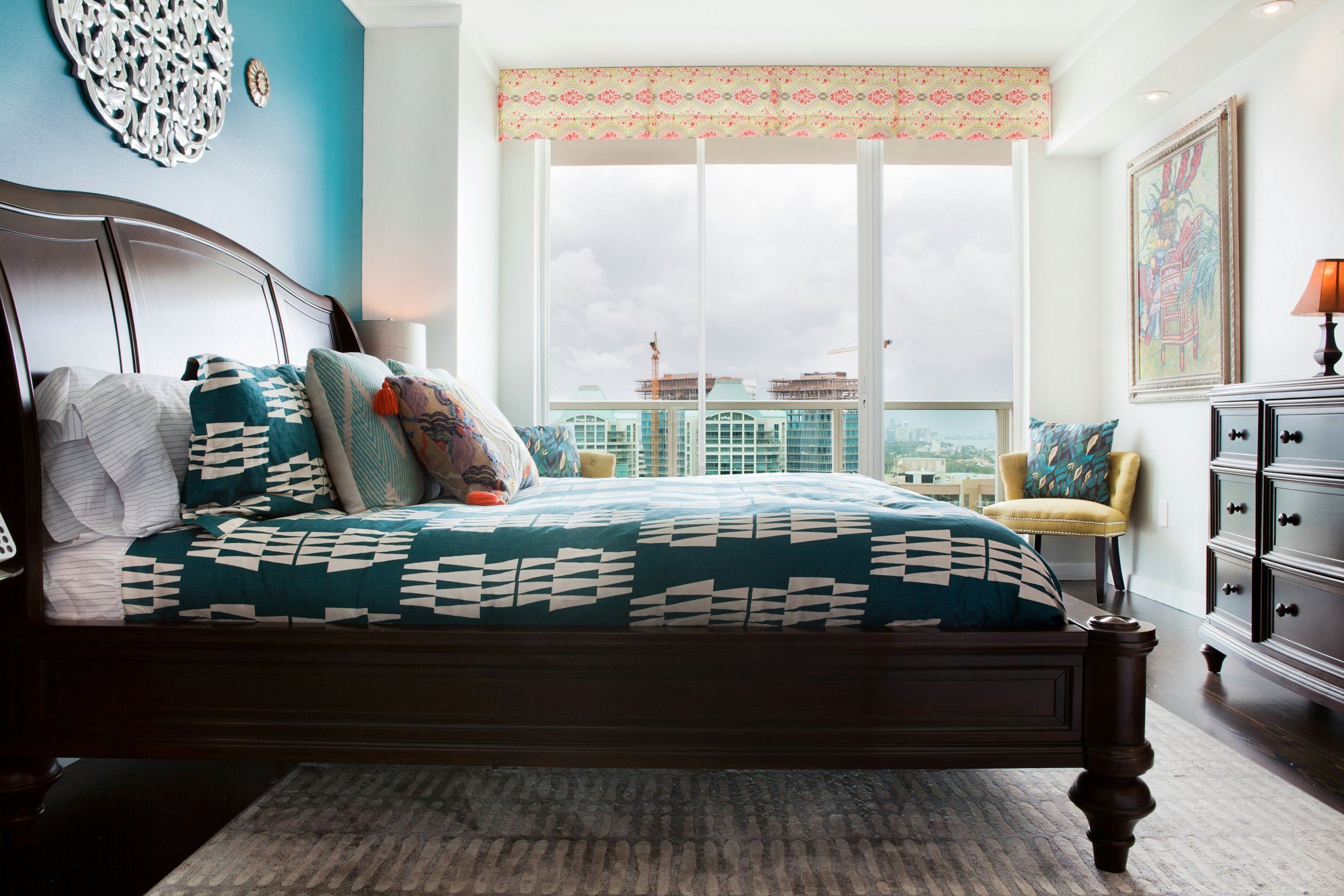 A bedroom with a large bed covered with a teal and white geometric-patterned quilt, multiple colorful pillows, dark wood furniture, a large window with city view, and artwork on the walls.