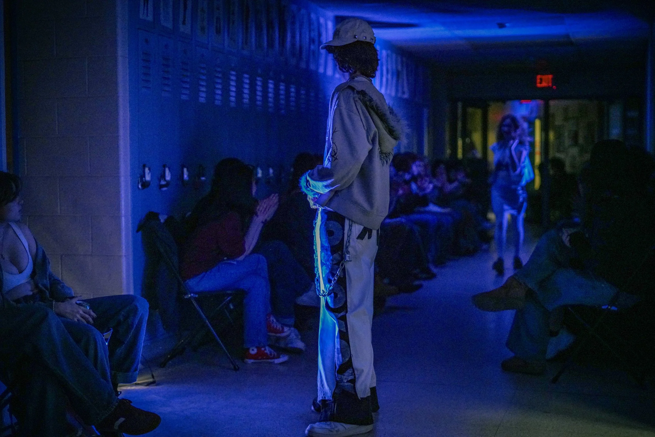 A person dressed in streetwear with chains, glasses, and a cap, standing in a dimly lit indoor space with blue lighting and other people in the background.