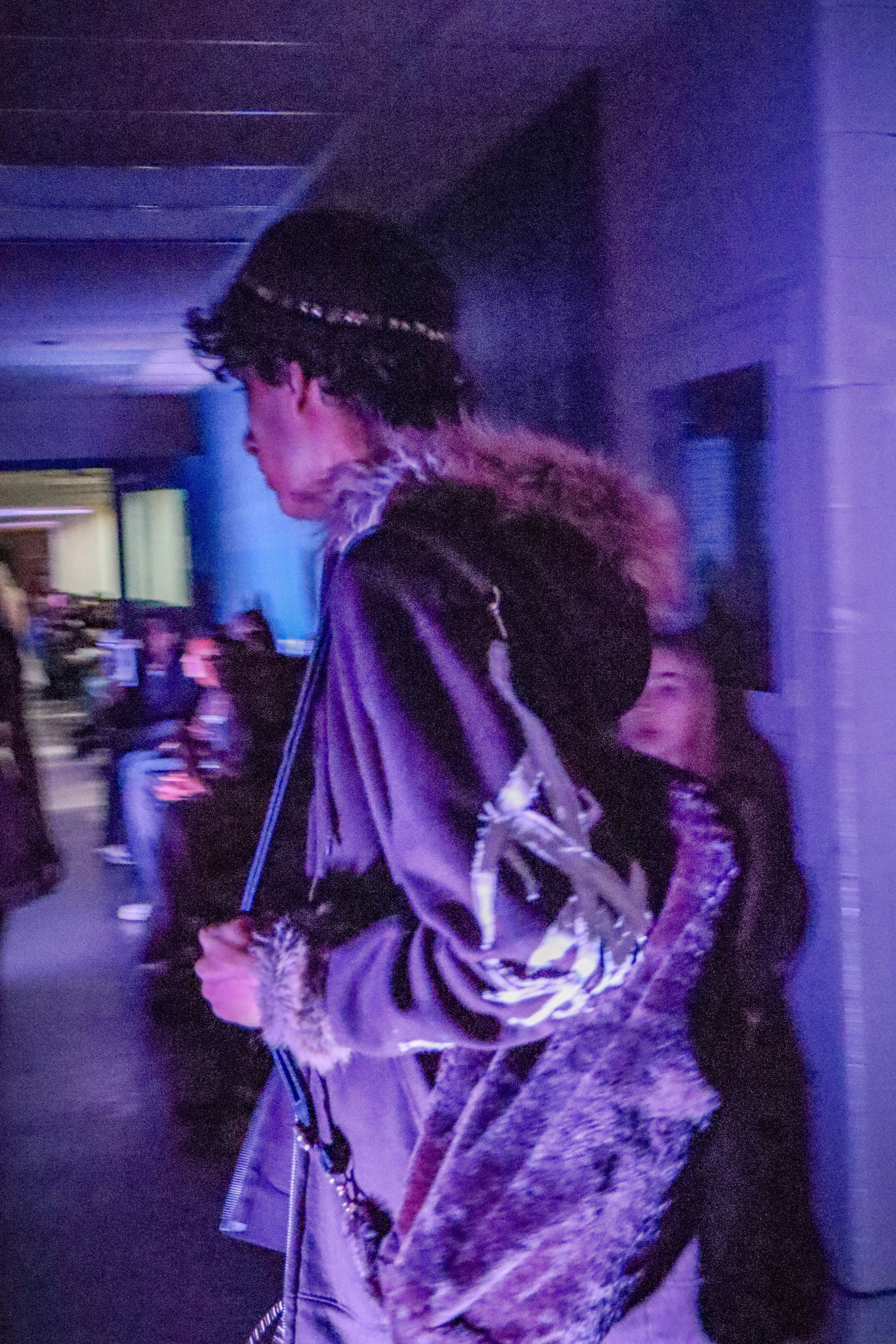 A person with curly hair wearing a black beanie with embellishments, a fur-lined jacket, and carrying a patterned backpack, walking in a dimly lit environment with purple lighting and blurred background.