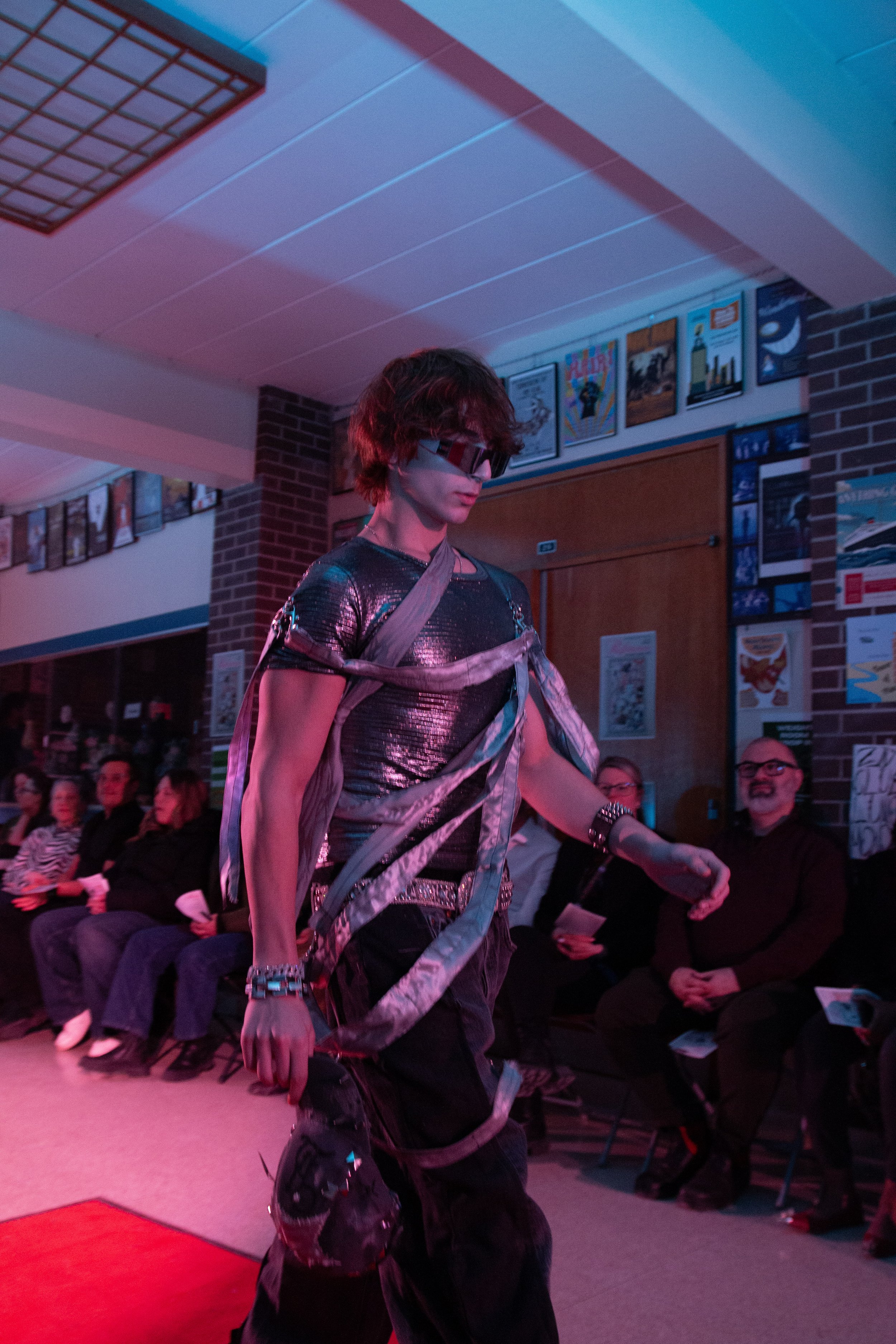 A person dressed in a shiny, metallic silver outfit with a matching hat, glasses, and gloves, walks on a runway in front of an audience, with purple lighting. Behind them are posters and a brick wall.