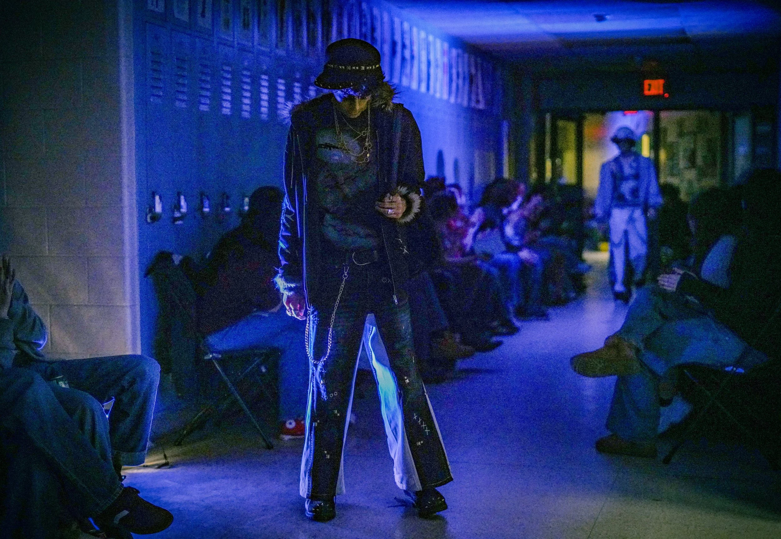 A person dressed in streetwear with chains, glasses, and a cap, standing in a dimly lit indoor space with blue lighting and other people in the background.