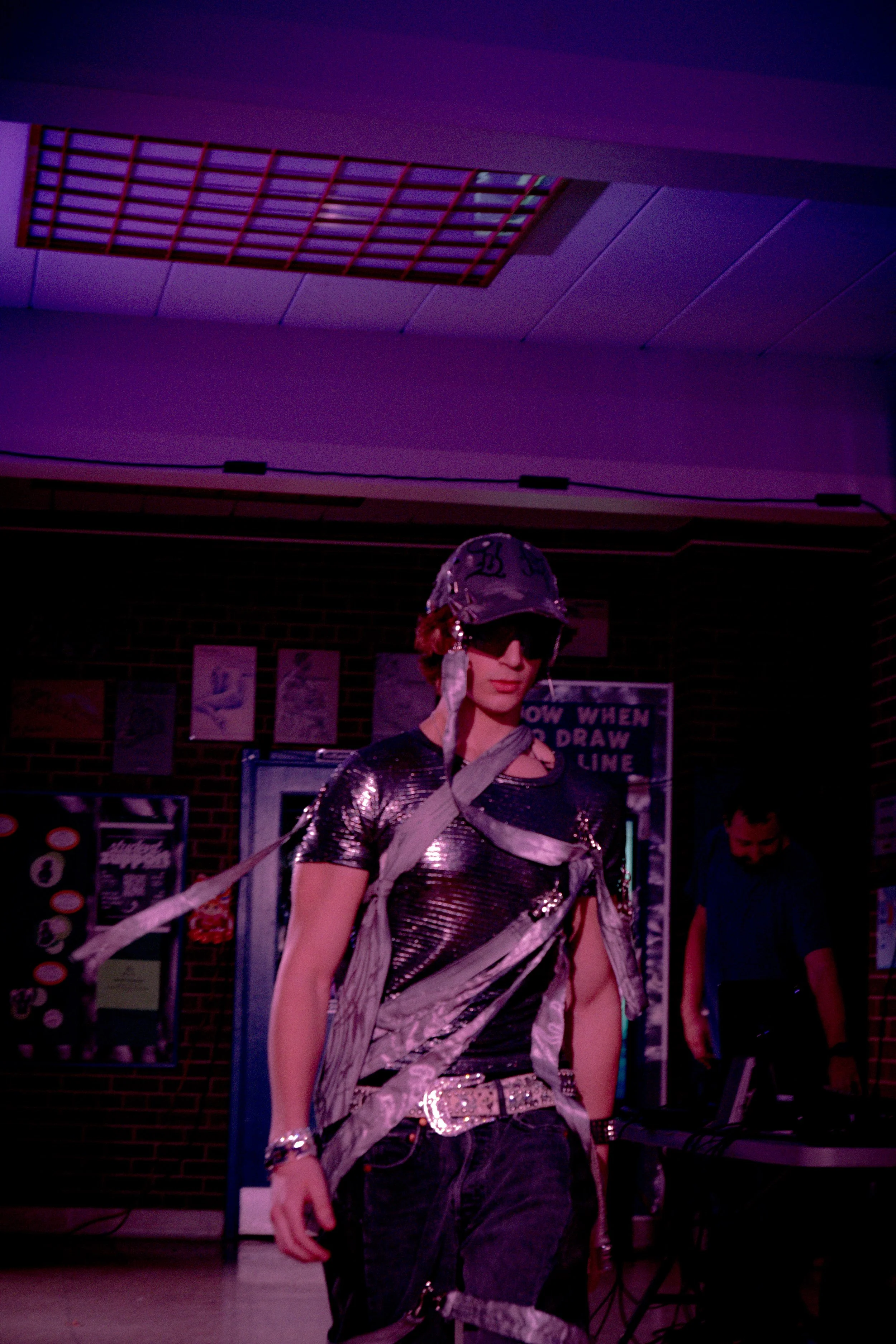 A person dressed in a shiny, metallic silver outfit with a matching hat, glasses, and gloves, walks on a runway in front of an audience, with purple lighting. Behind them are posters and a brick wall.