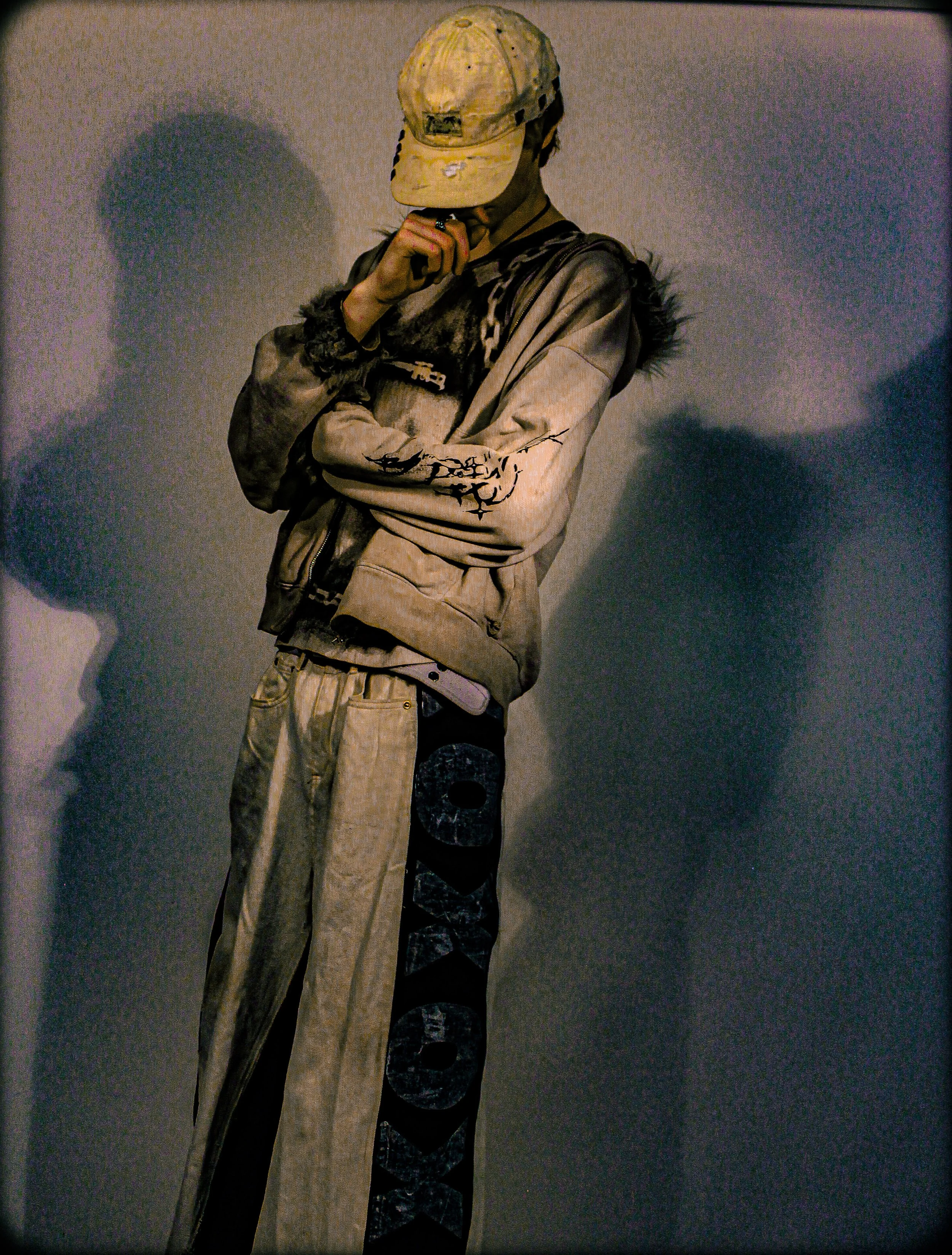A person in a beige jacket, patterned pants, and a yellow cap with a worn logo, standing against a dark wall with their head bowed and hand near their chin.