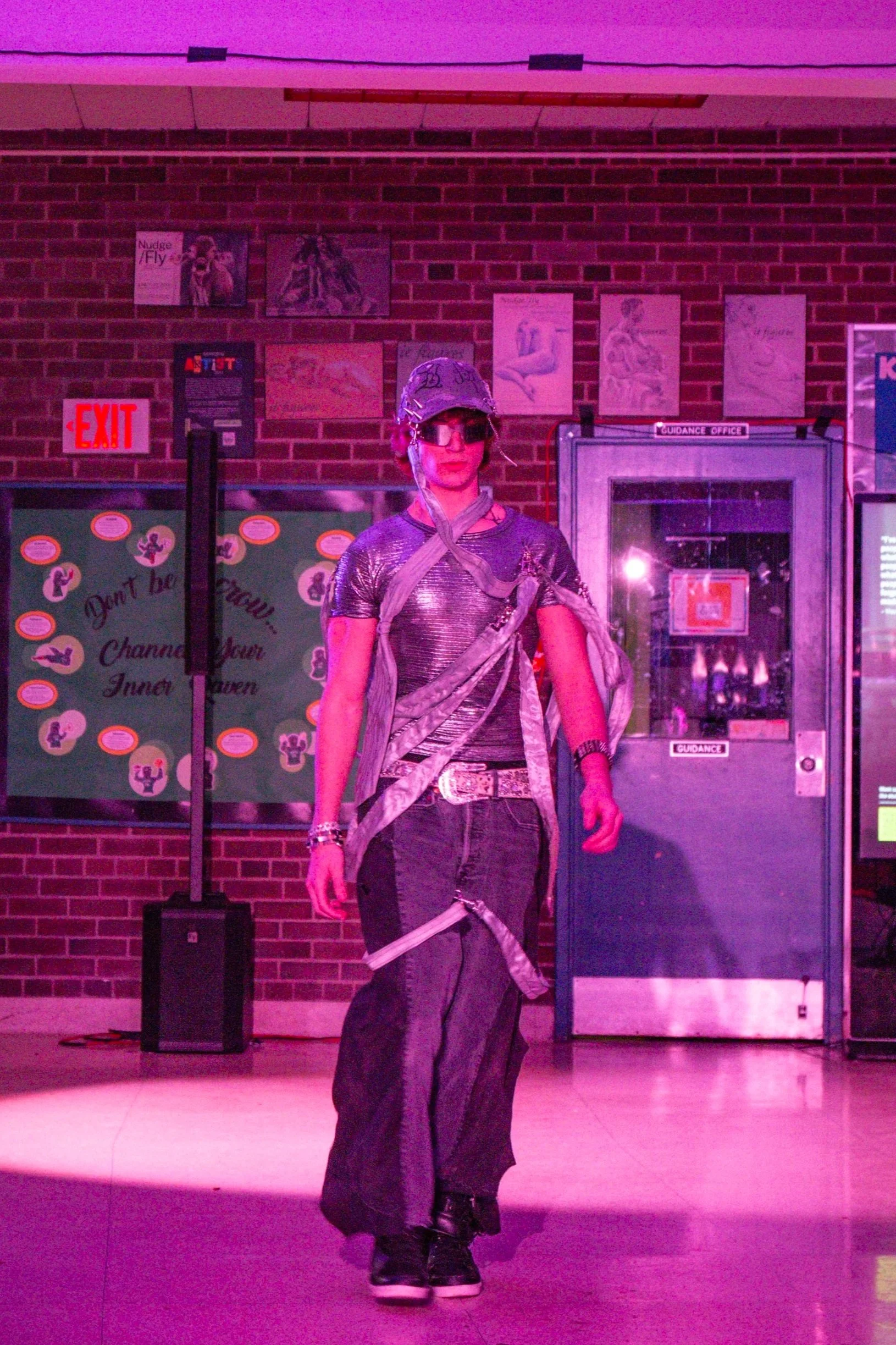 Person dressed in dark, shiny, metallic clothing with a helmet and sunglasses, standing in front of a brick wall with posters, exit sign, and a guidance office door, illuminated by purple lighting.