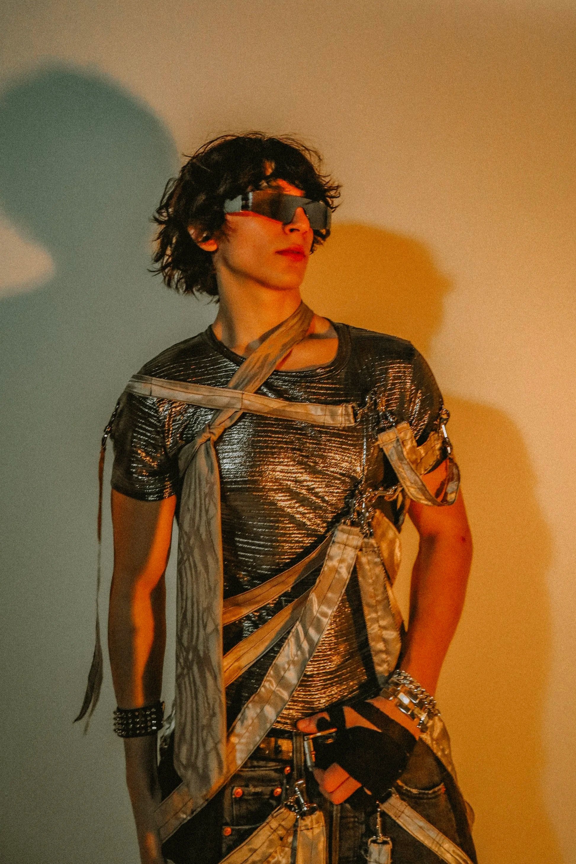 A person with curly dark hair wearing large futuristic silver reflective sunglasses, a metallic silver textured shirt, and a layered outfit with straps, chains, and accessories, standing against a plain background with yellow and blue lighting.