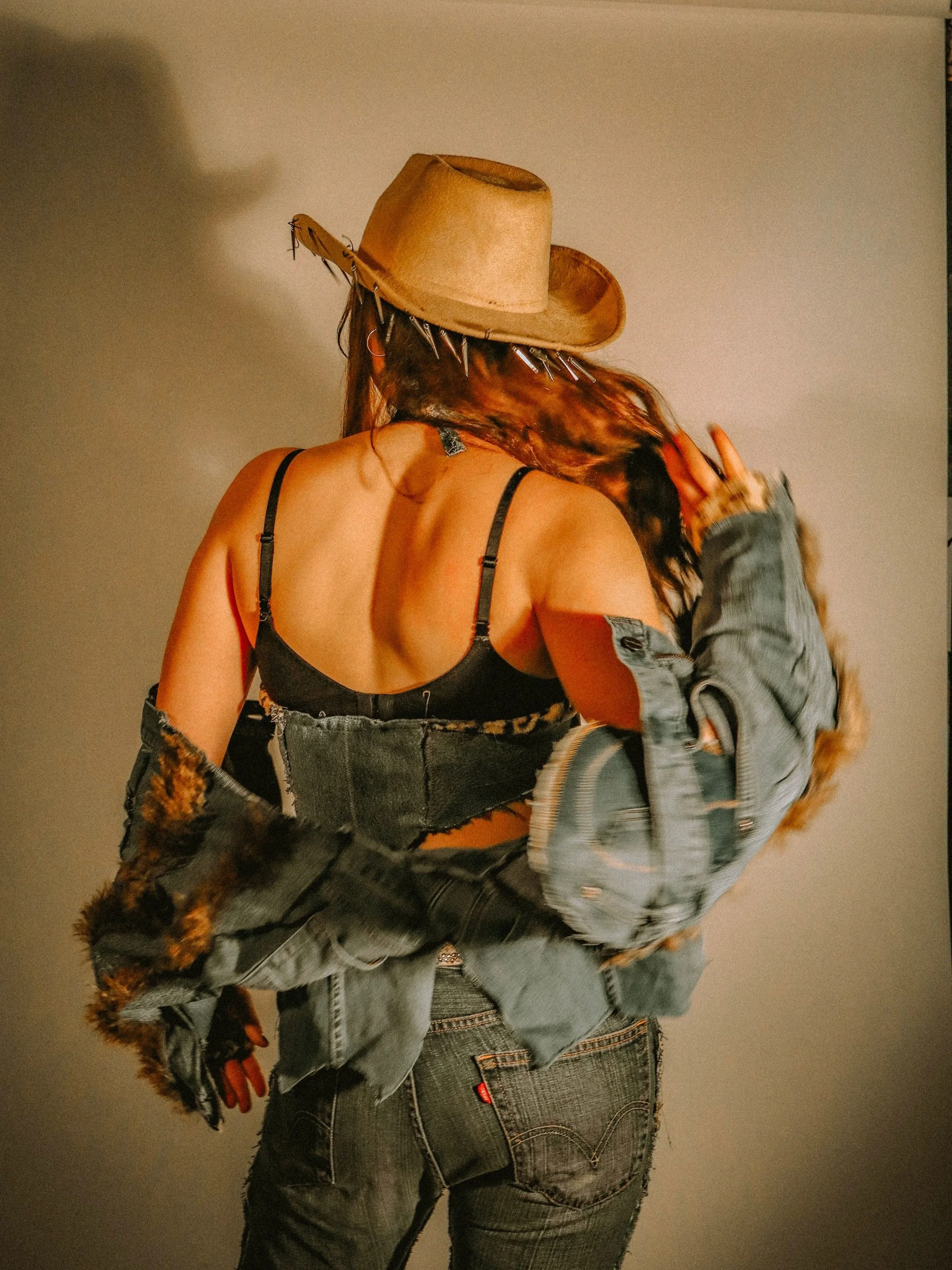 Person with long hair wearing a tan hat with spikes, a black tank top, a denim jacket with fur, and ripped jeans, standing with back facing the camera against a plain wall.