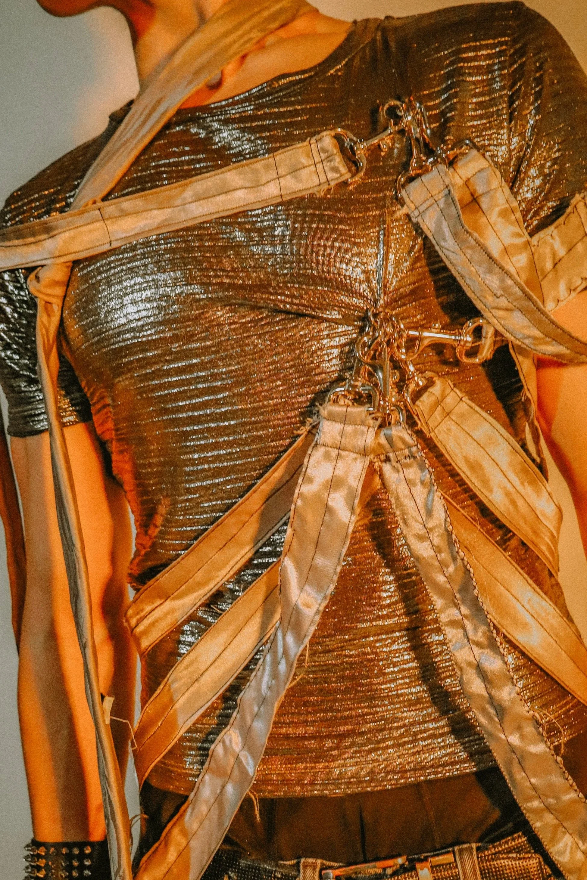 Close-up of a person wearing a shiny, metallic, pleather shirt with a belt and many straps, along with accessories like a studded bracelet.