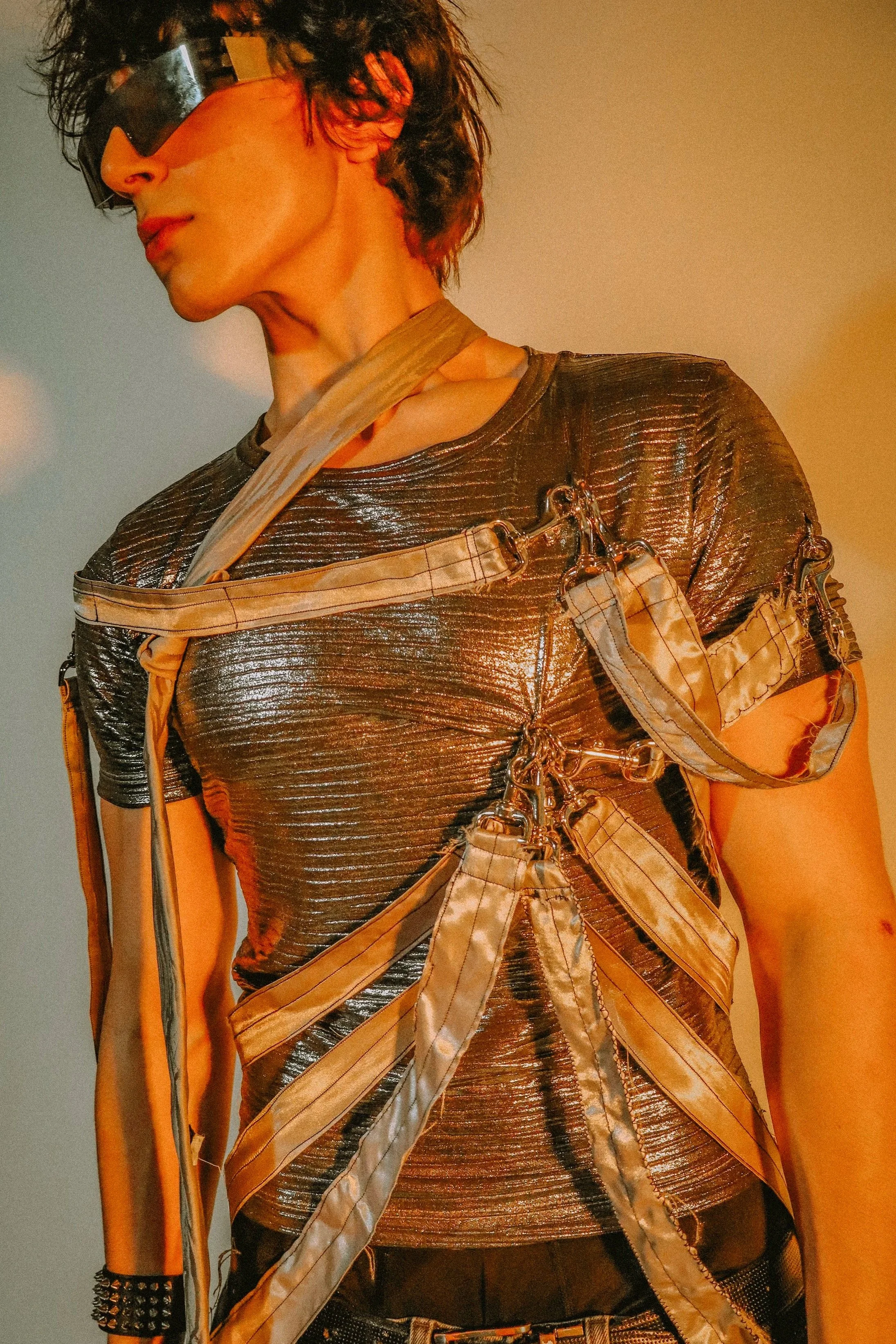 A person with curly dark hair wearing large futuristic silver reflective sunglasses, a metallic silver textured shirt, and a layered outfit with straps, chains, and accessories, standing against a plain background with yellow and blue lighting.