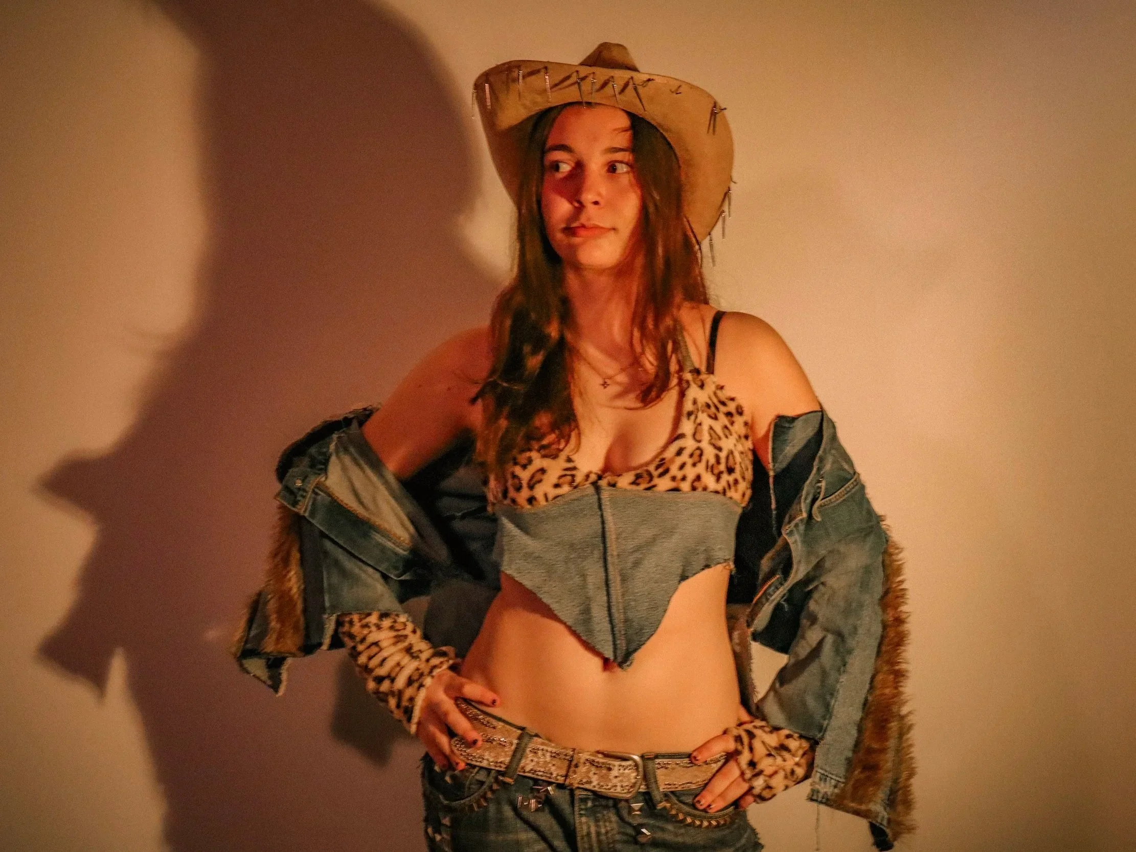 A young woman wearing a denim-leopard print top, denim-fur jacket, and a cowboy hat with spikes, standing against a plain wall.