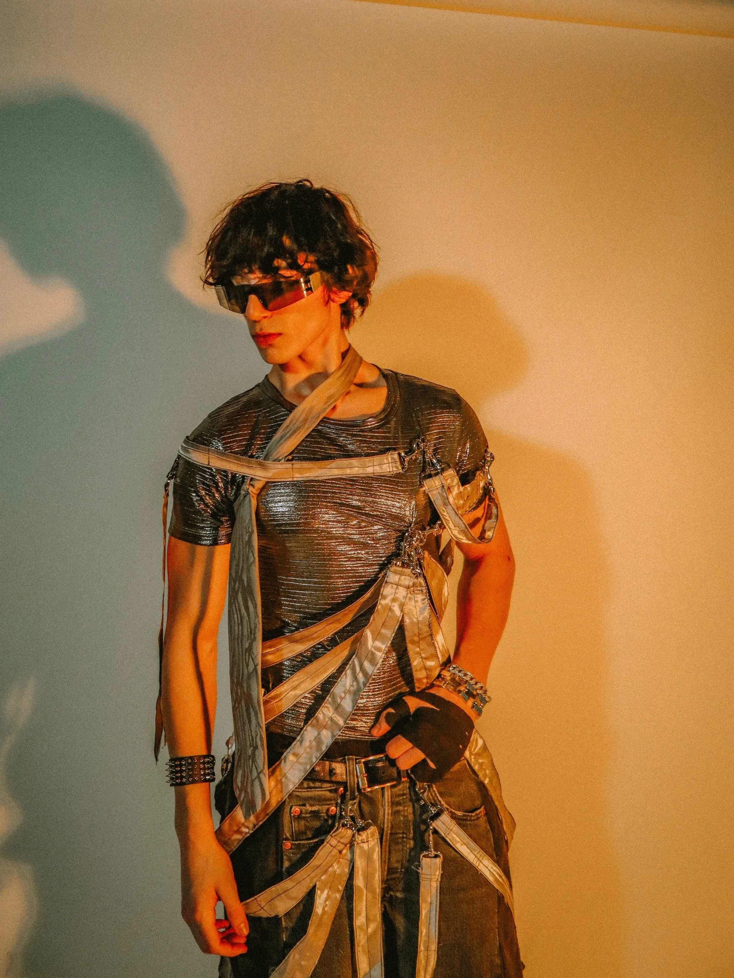 A person with curly dark hair wearing large futuristic silver reflective sunglasses, a metallic silver textured shirt, and a layered outfit with straps, chains, and accessories, standing against a plain background with yellow and blue lighting.