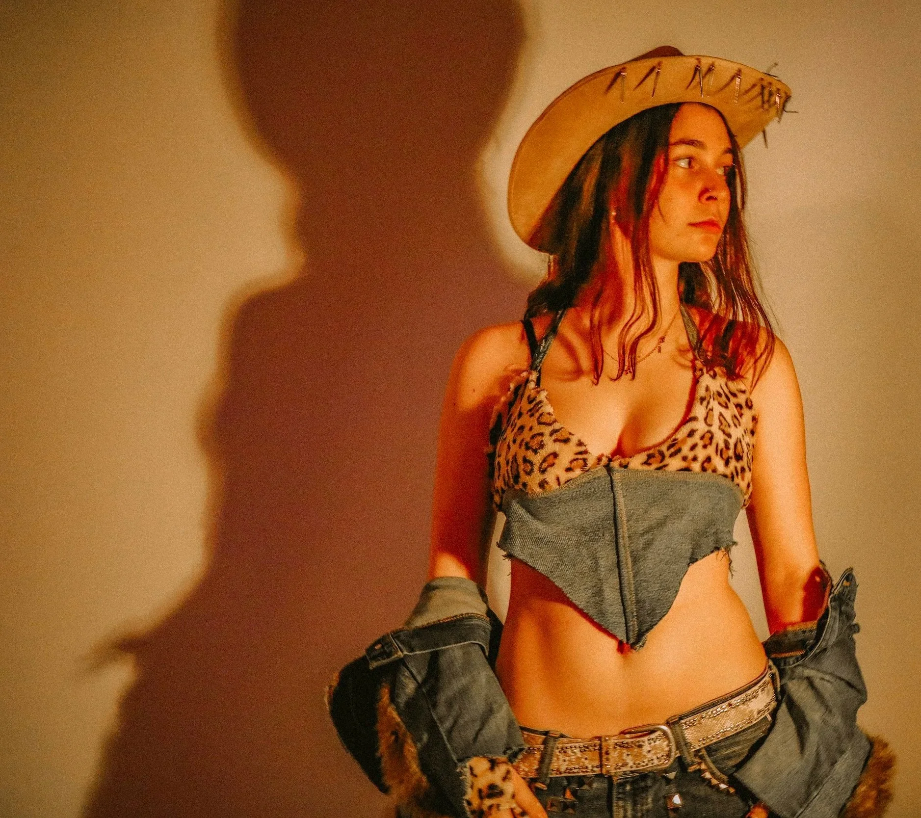 Young woman wearing a leopard print crop top, gray shorts, leather belt, denim jacket hanging off her arms, wide-brimmed straw hat with embellishments, and jewelry, standing against a light-colored background.
