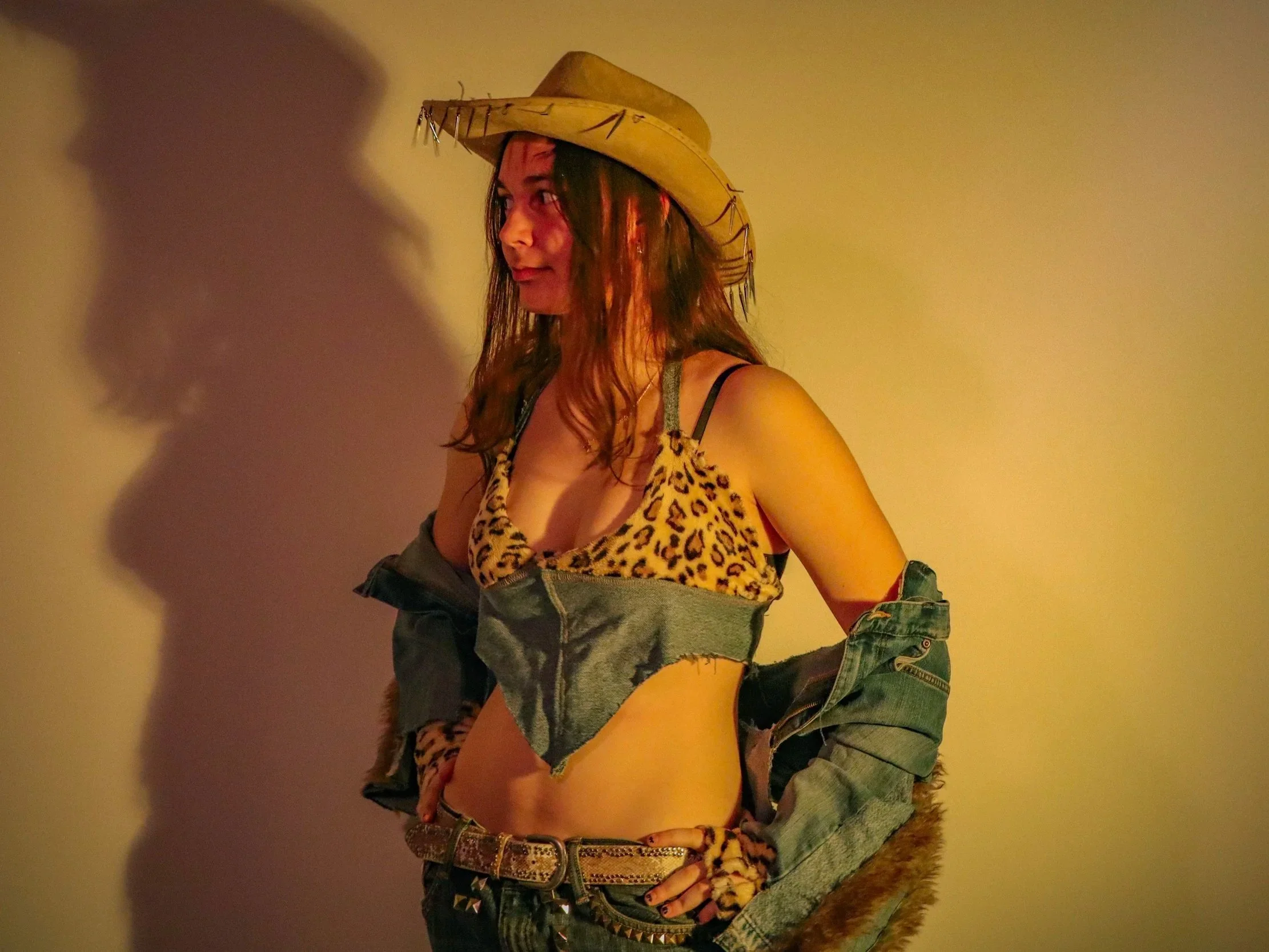 A woman wearing a leopard print top, denim shorts, and a large straw hat with decorative accents, standing against a plain wall with her right hand on her hip.